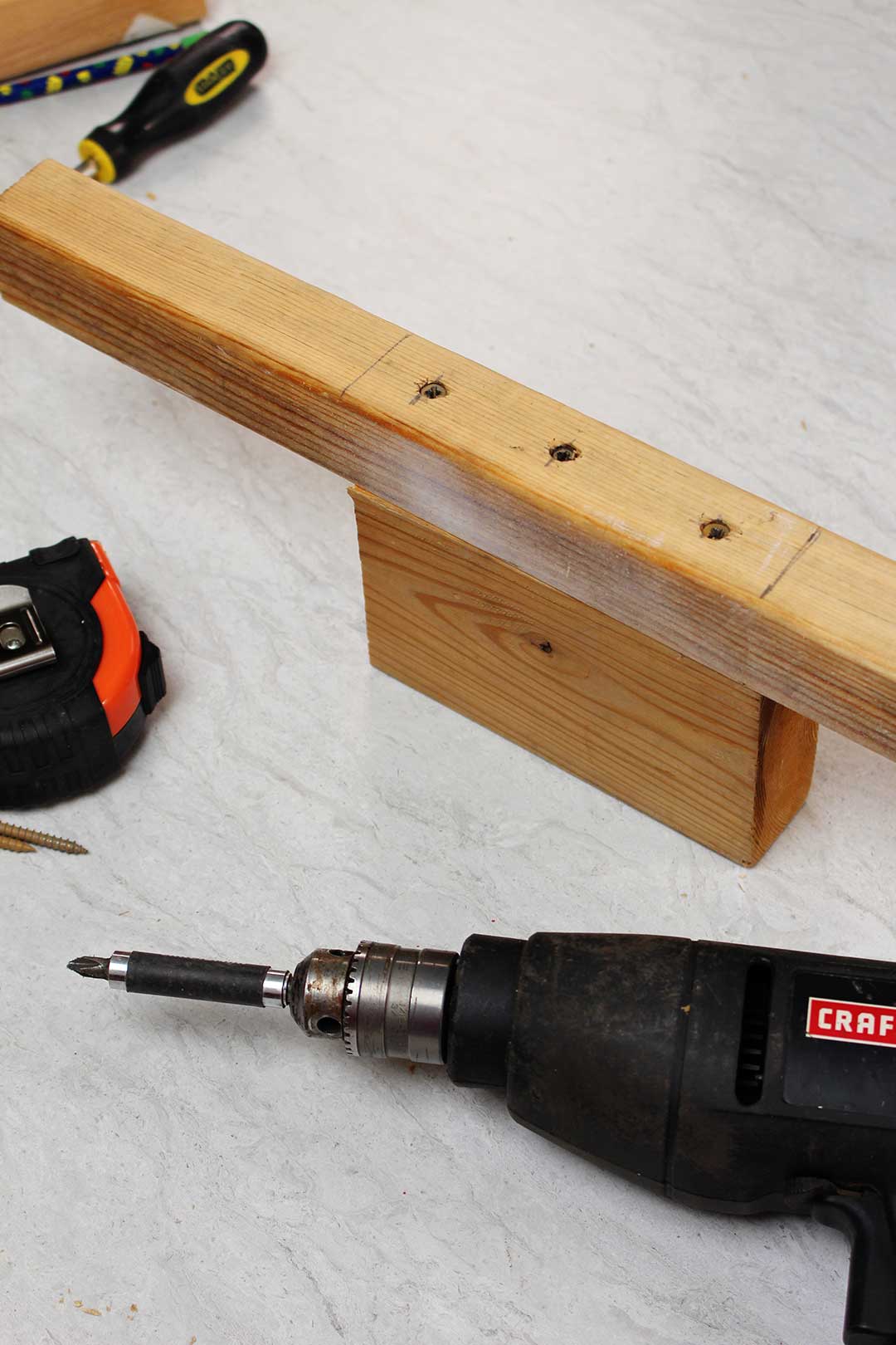 Close up image of drill, measuring tape and two pieces of wood screwed together for stilts.