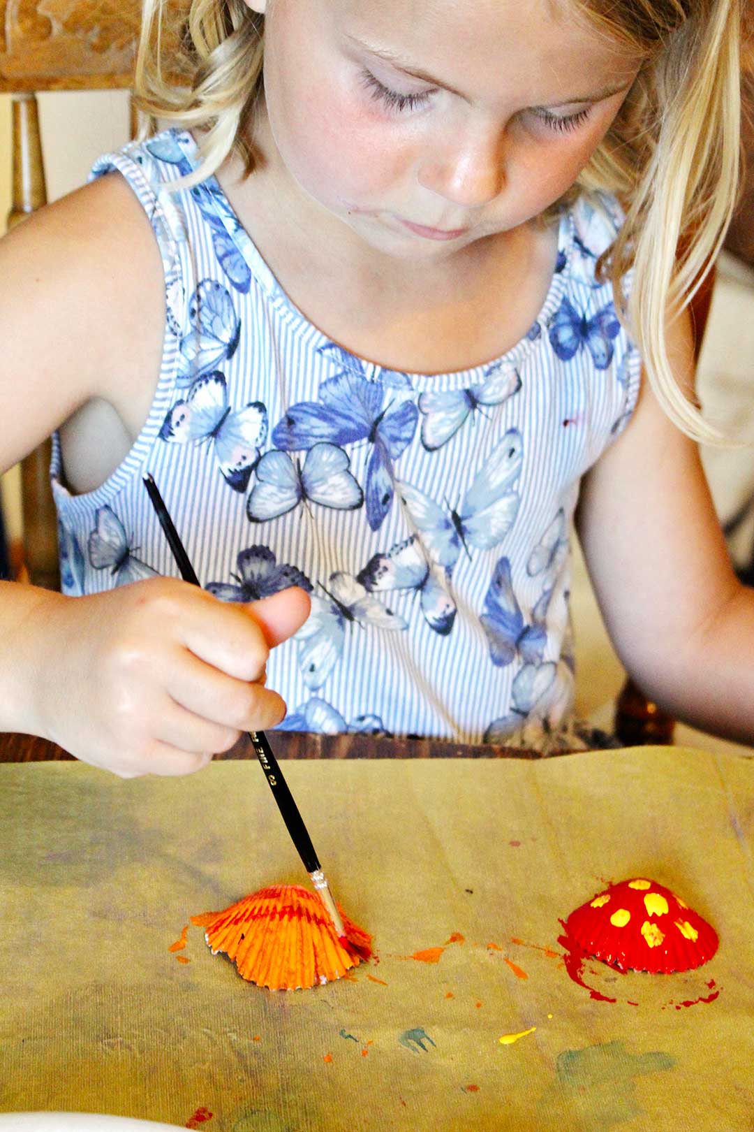 Blonde girl with blue butterfly dress paints seashells on yellow table cloth.