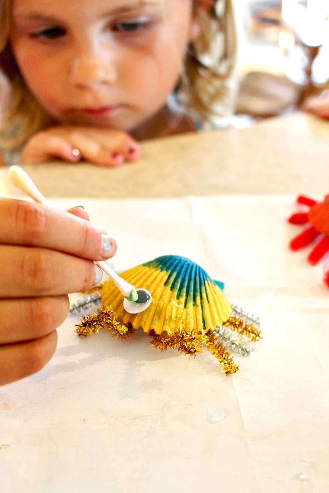 Small girl watches person place googly eye on yellow painted shell crab.