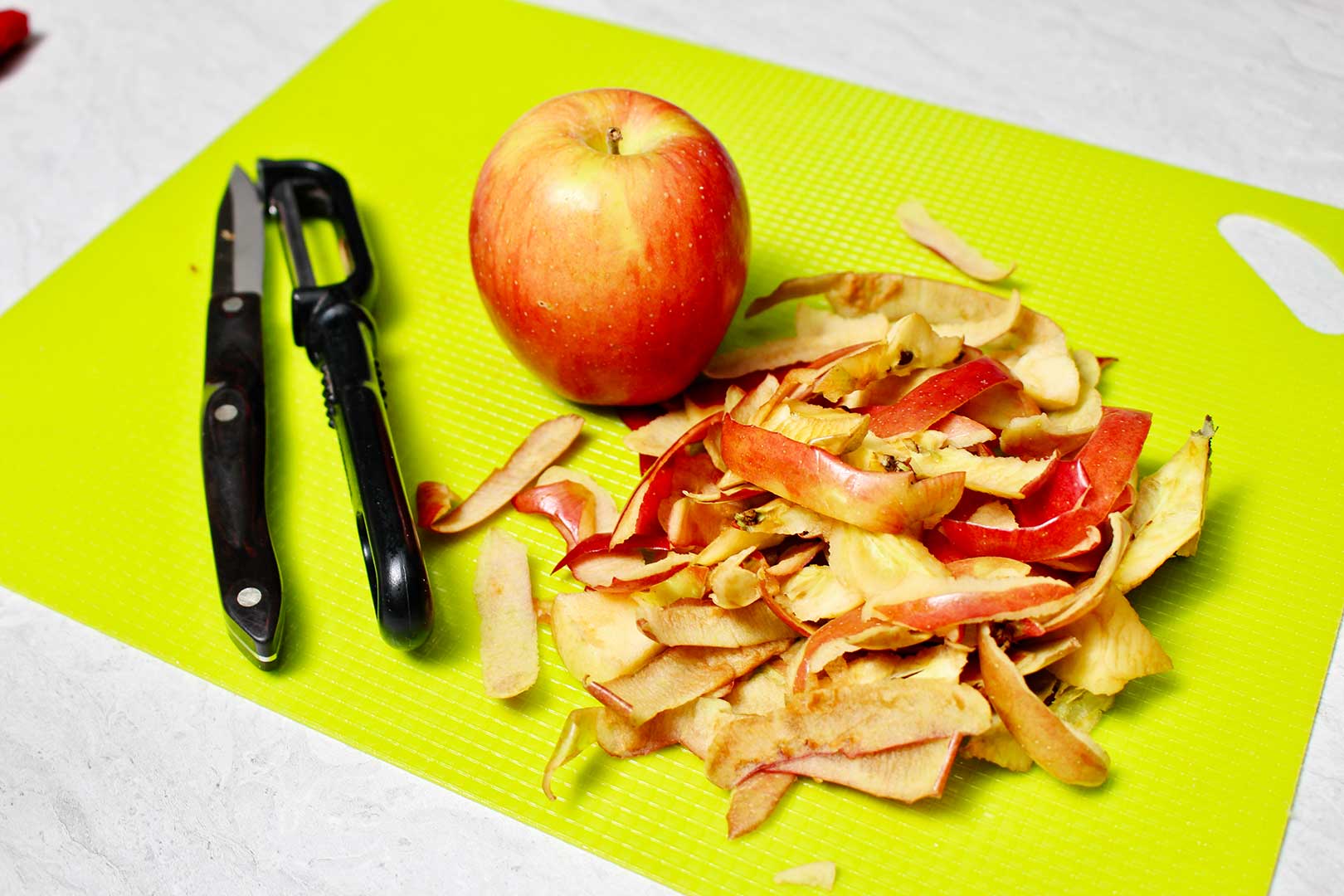 Apple and apple peels on a neon green plastic cutting board with a knife and peeler next to them.