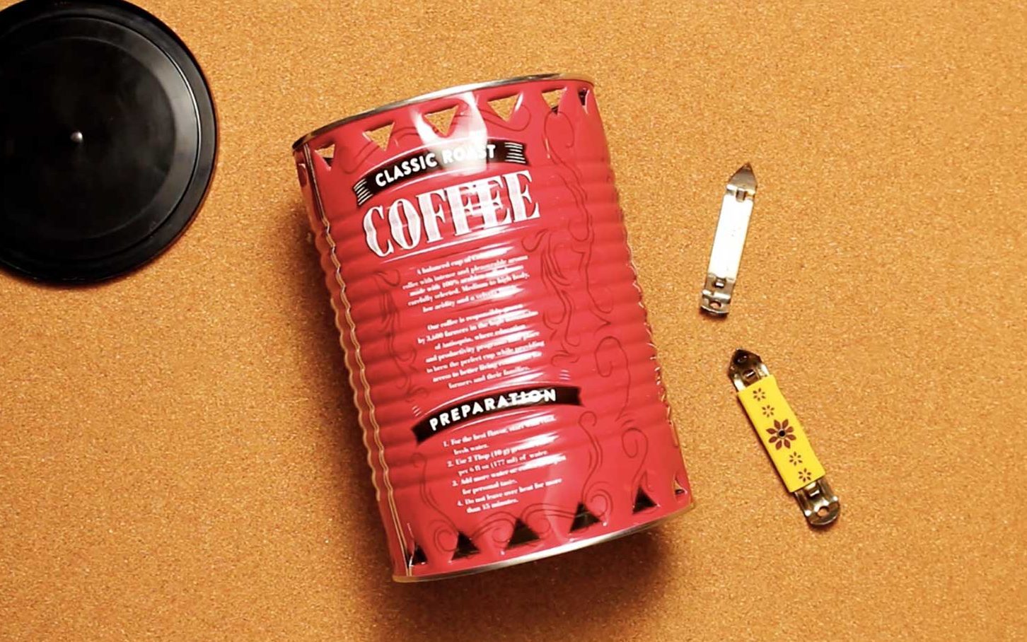 A coffee can, lid and two small can openers resting on cork colored background.