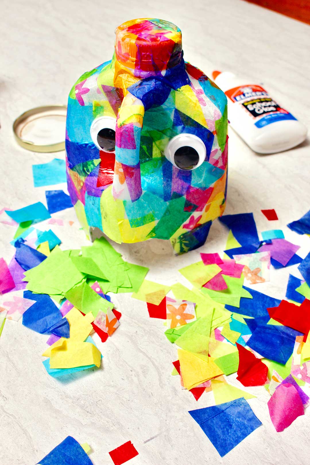 Semi complete elephant craft with many colorful pieces of cut up tissue paper and glue resting near the project.