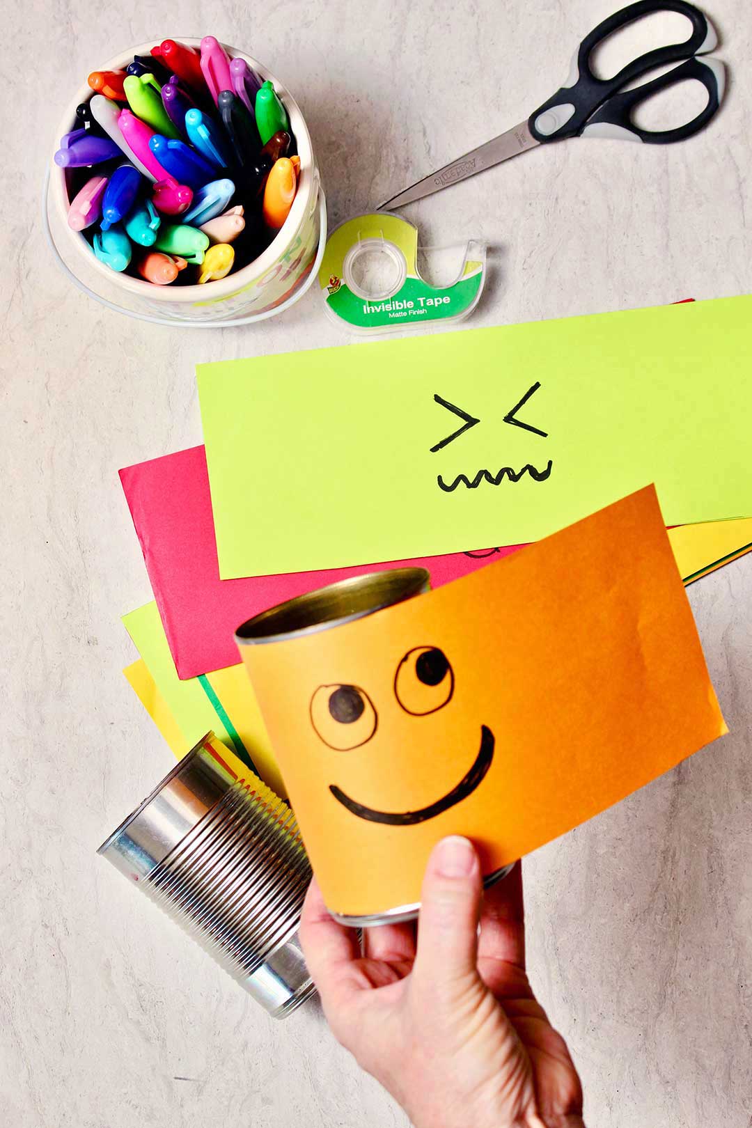 A hand wrapping an orange strip of paper with a smiley face drawn on it to a tin can with other supplies in the background.