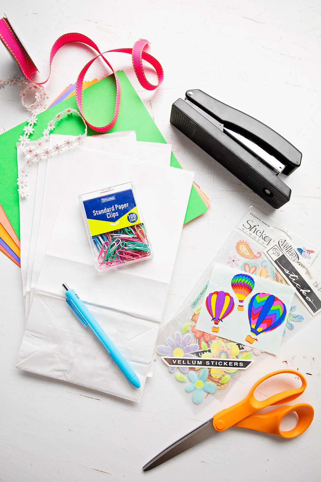 Supplies for a paper bag book. Stapler, scissors, stickers, pen, colorful paper clips, white paper bags, colorful paper and pink ribbon.
