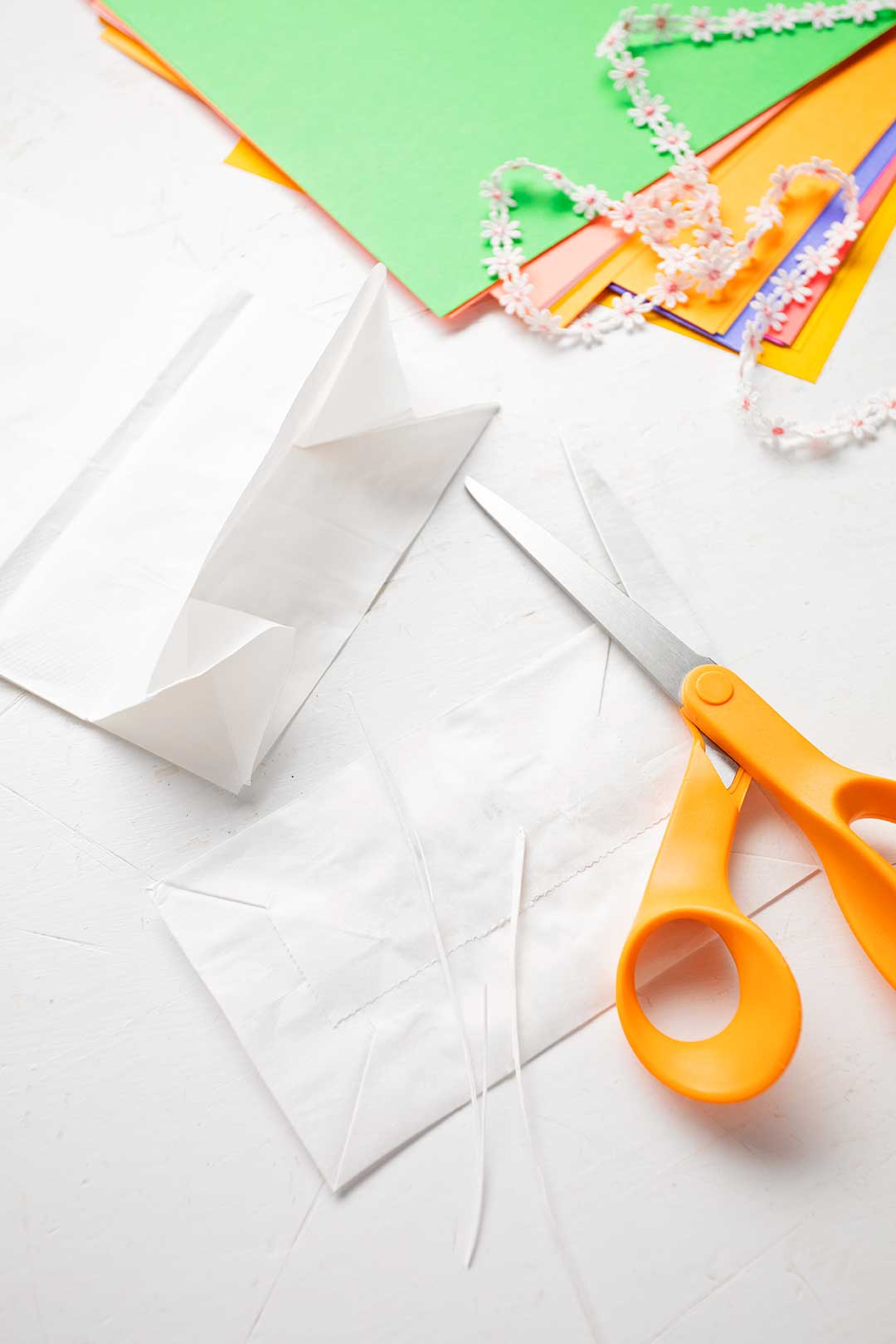 Scissors and colorful paper rest on a white background. The image shows the bottom of a white paper bag cut off.