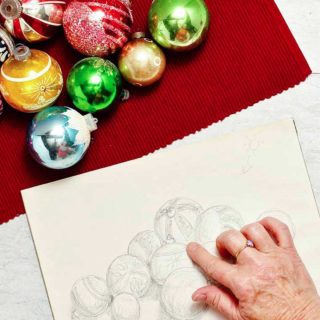 Christmas ornaments lay on a red placemat. A hand holds a red pencil and shades finishing touches on the drawing.
