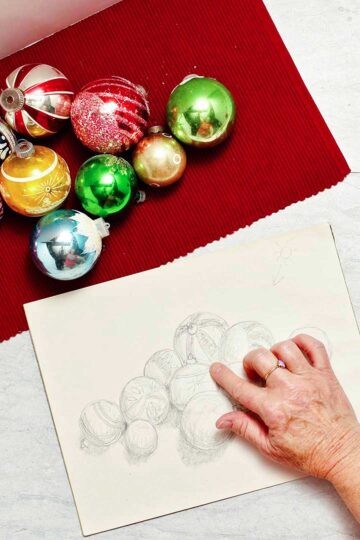 Christmas ornaments lay on a red placemat. A hand holds a red pencil and shades finishing touches on the drawing.