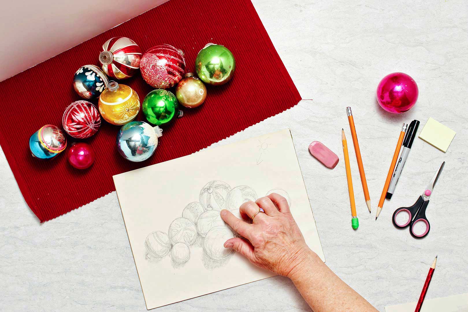 Christmas ornaments lay on a red placemat and person blends some of their drawing with their finger.