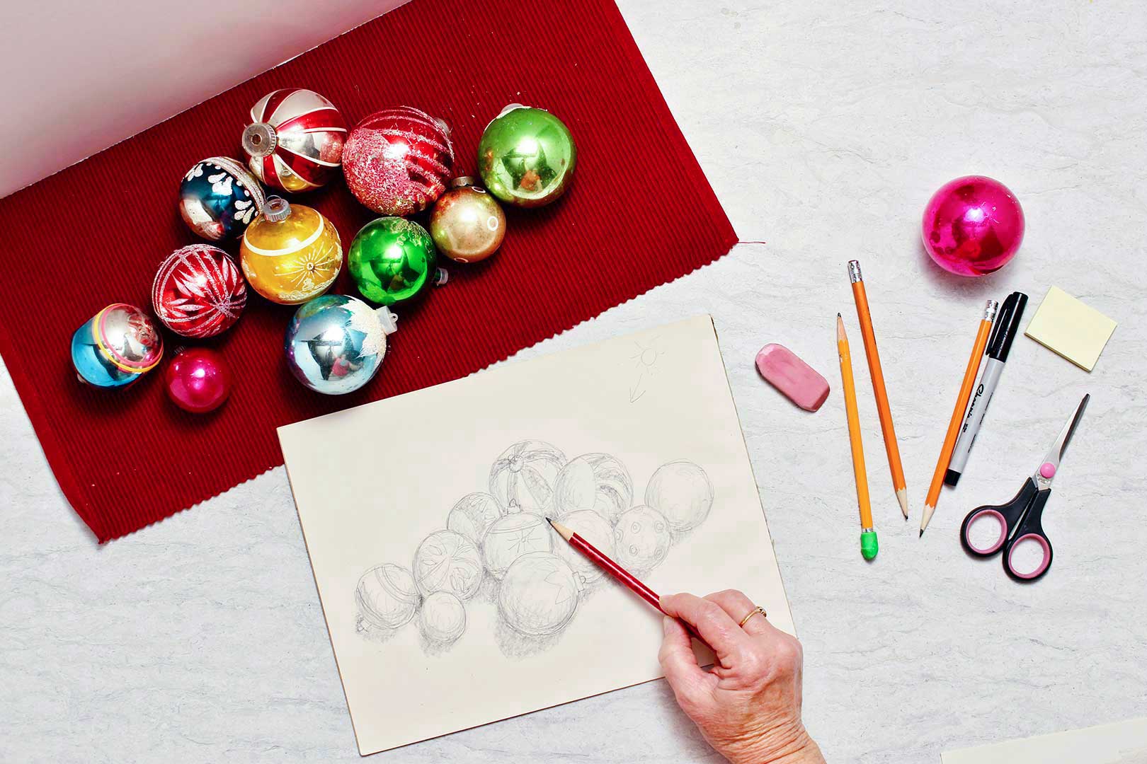 Christmas ornaments lay on a red placemat. A hand holds a red pencil and shades finishing touches on the drawing.
