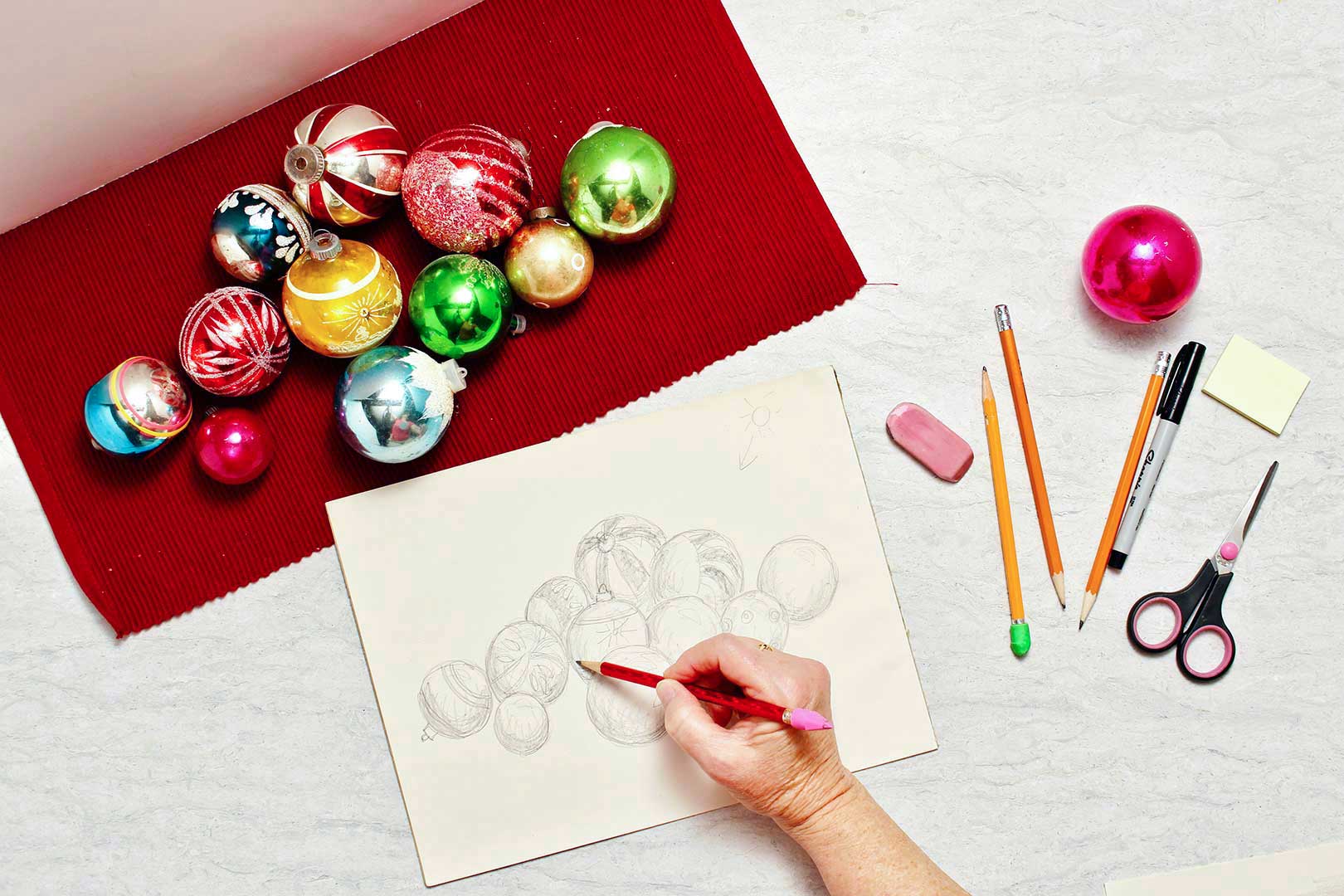 Christmas ornaments lay on a red placemat and a person puts finishing touches on their Christmas ornament pencil drawing.