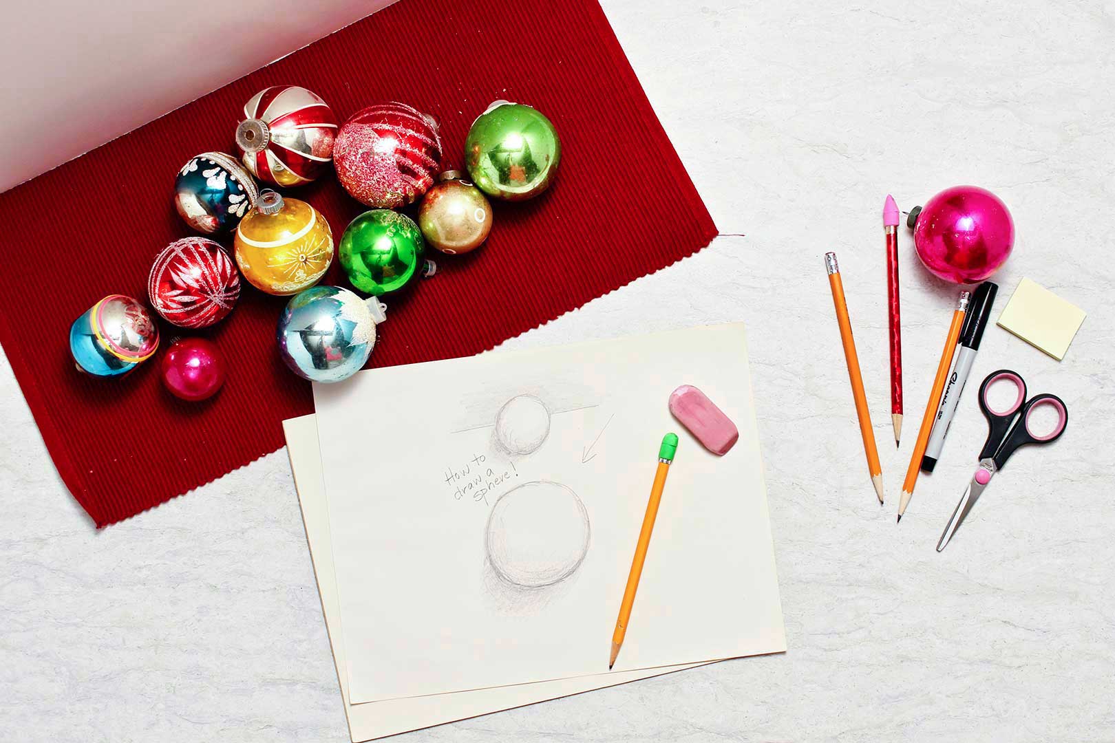 Christmas ornaments lay on a red placemat and a drawing of two spheres rests near by with a pencil and eraser on top of it.
