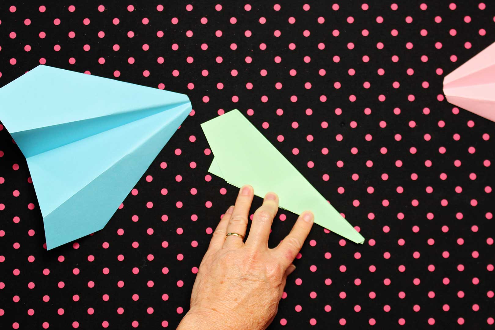 A hand showing individual folds instructing how to make a paper airplane out of green paper. Two finished planes sit next to it on a black and pink polka dotted background.