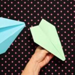 A hand showing individual folds instructing how to make a paper airplane out of green paper. Two finished planes sit next to it on a black and pink polka dotted background.