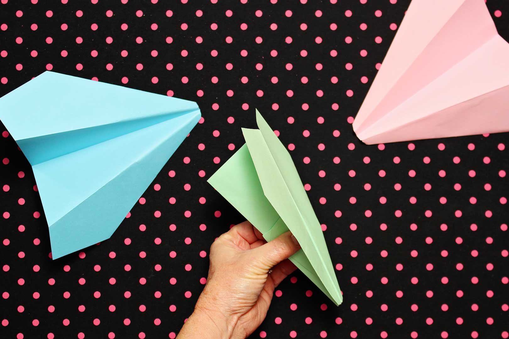 A hand showing individual folds instructing how to make a paper airplane out of green paper. Two finished planes sit next to it on a black and pink polka dotted background.