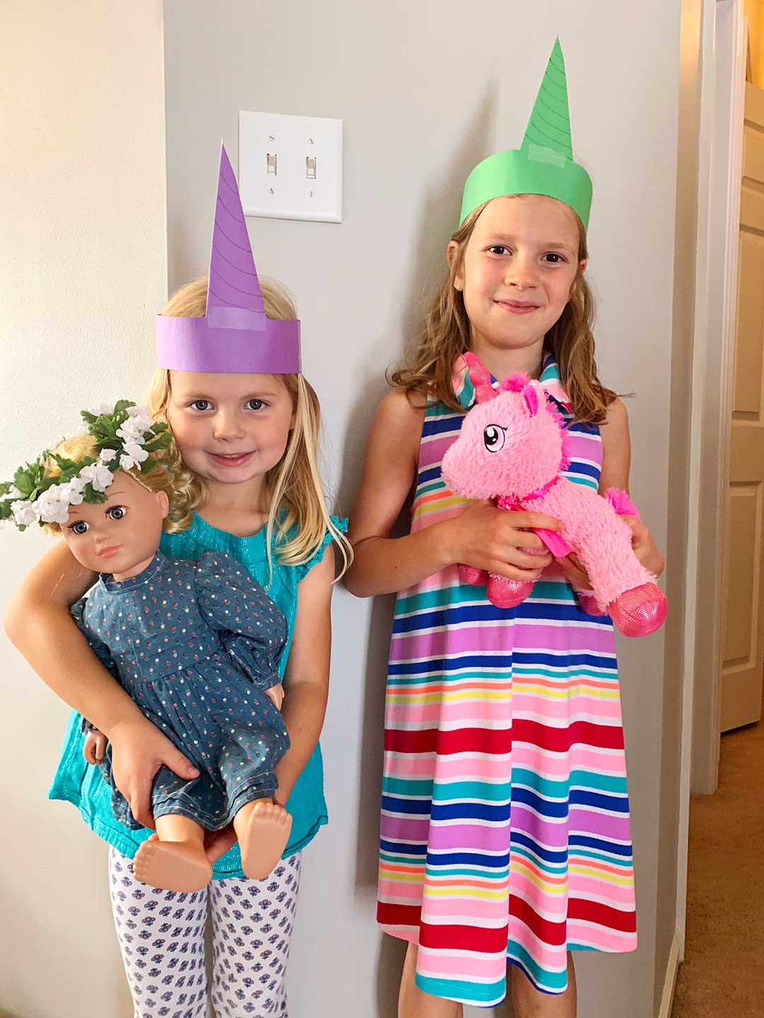 Two girls wearing unicorn crowns smile at camera holding a doll and a unicorn stuffed animal.