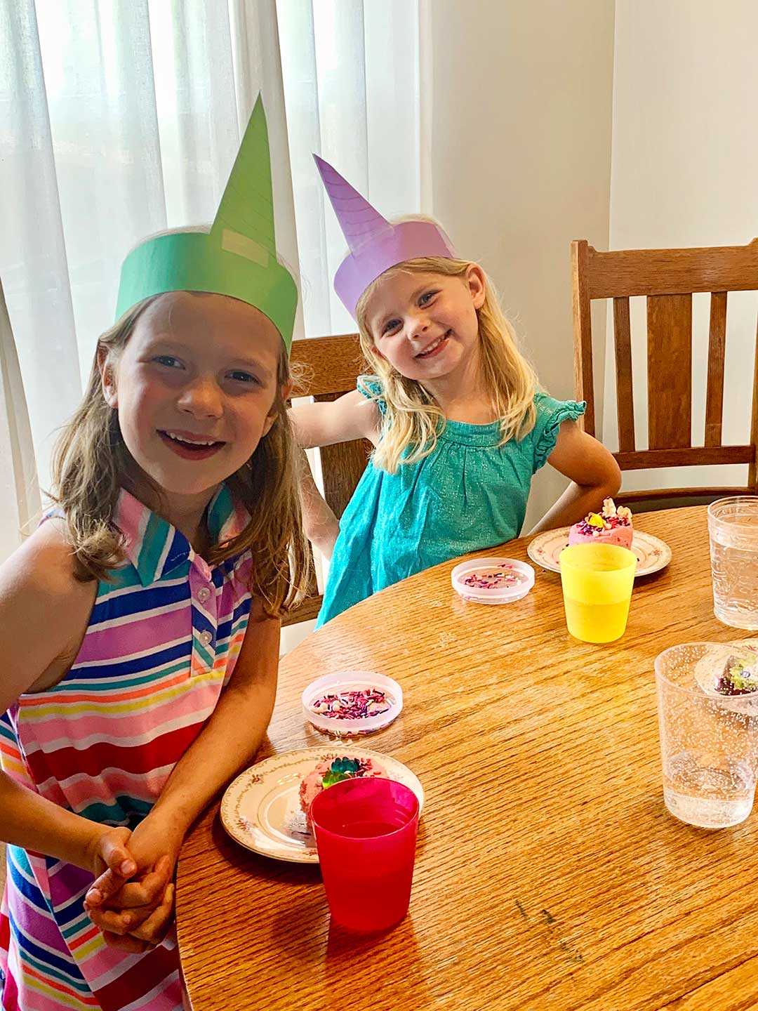 Two young girls wearing unicorn crowns made from paper sit at a kitchen table decorating cupcakes with sprinkles.
