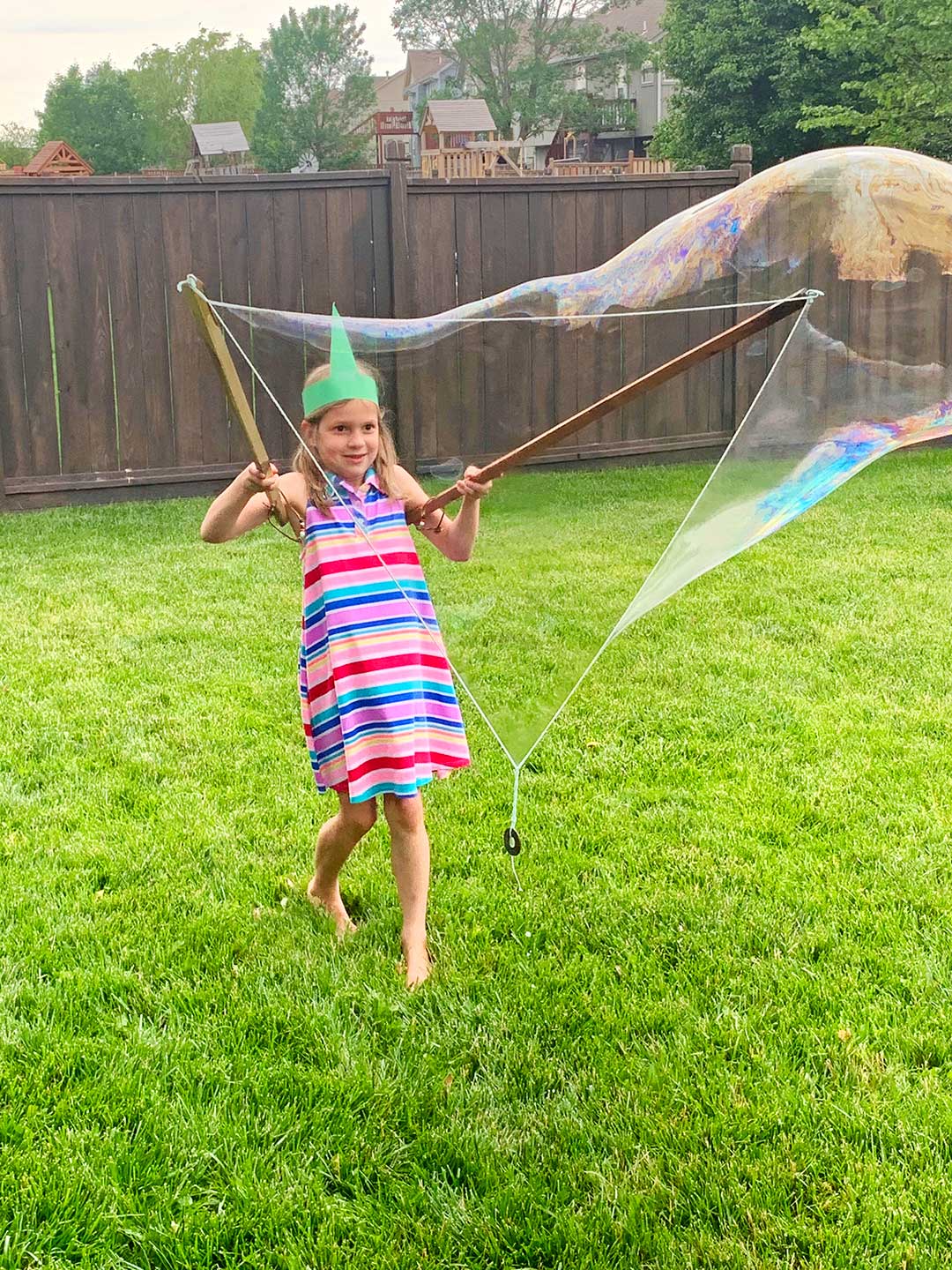 Young girl with striped dress and unicorn crown plays with large bubble wand in the back yard.