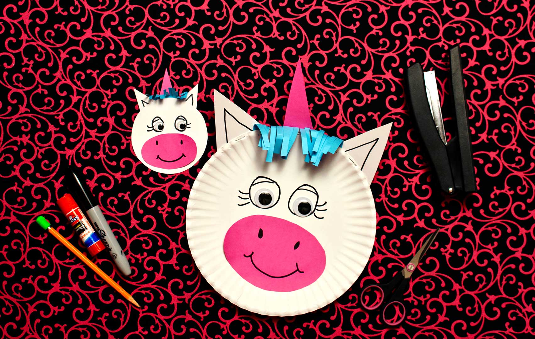 Paper plate unicorn head rest on decorative neon pink and black background with scissors, stapler, pencil, glue stick and sharpie.