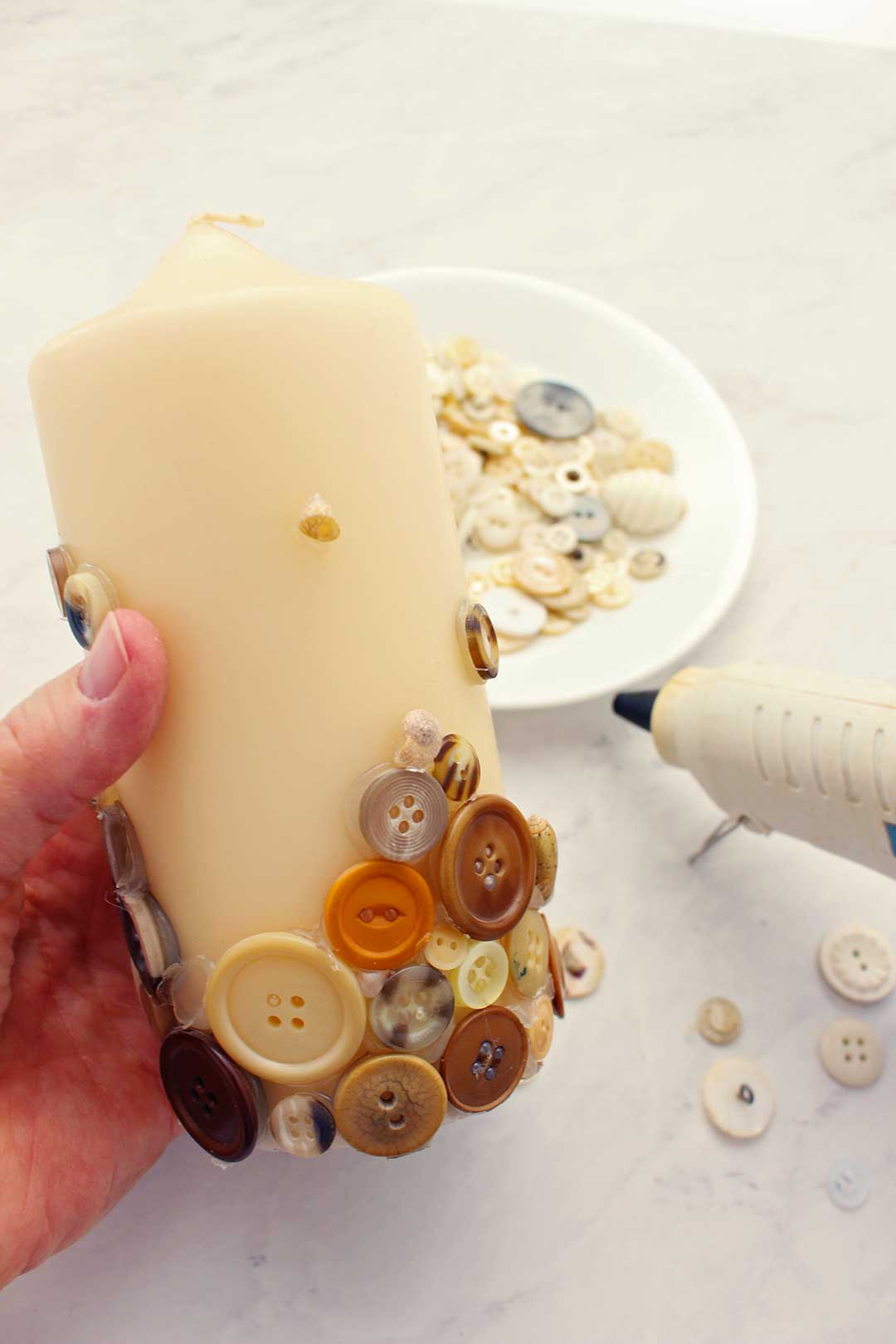 Hand holding off white pillar candle with tan buttons hot glued on the bottom.