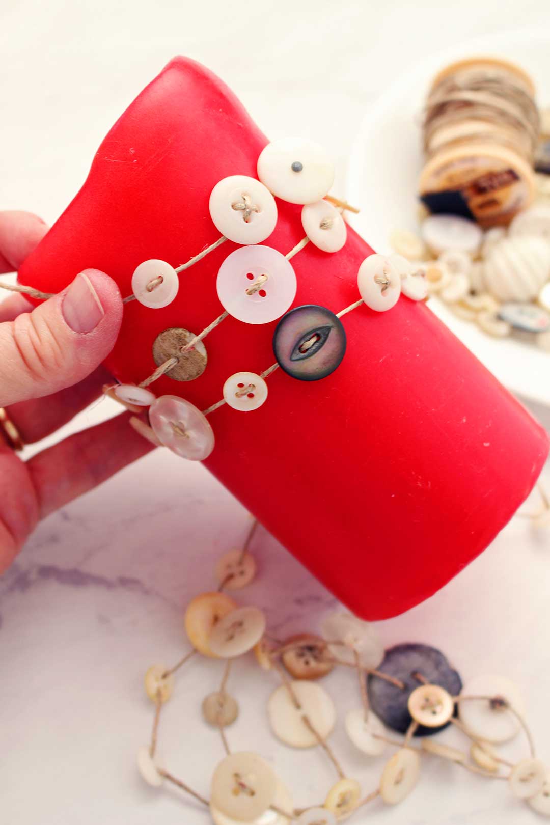 Hand holding red pillar candle wrapped with buttons and twine.