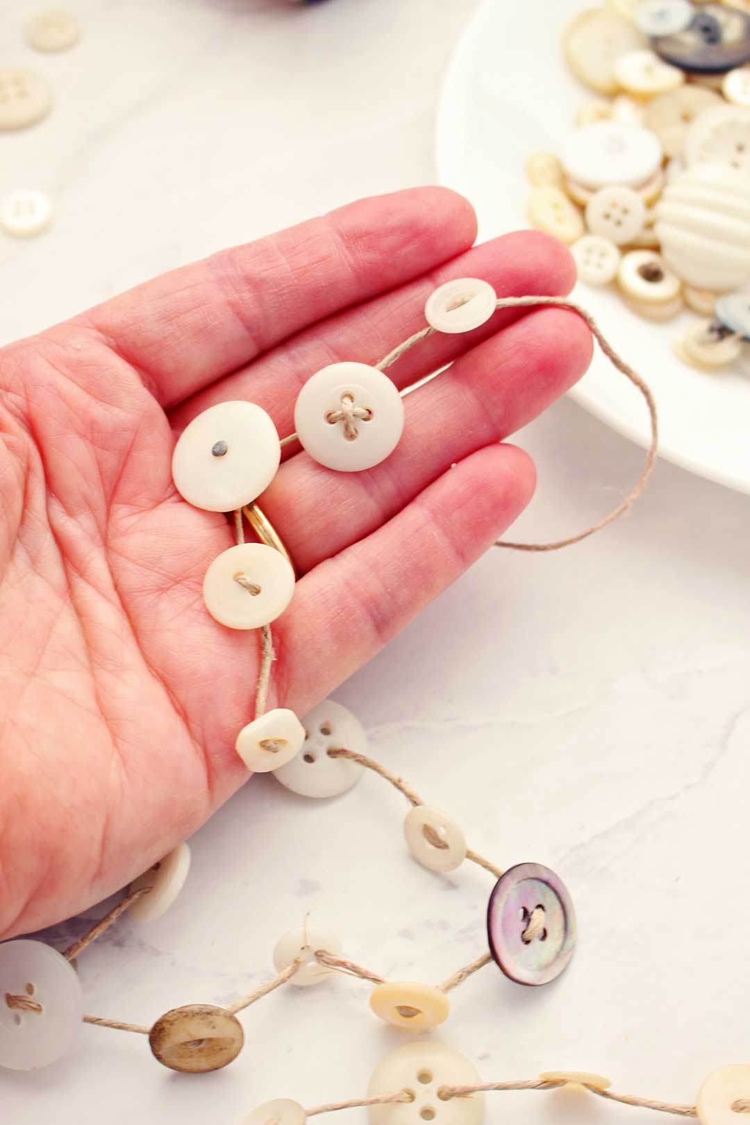 Open hand holding string of twine and off white colored buttons with plate of buttons in the background.