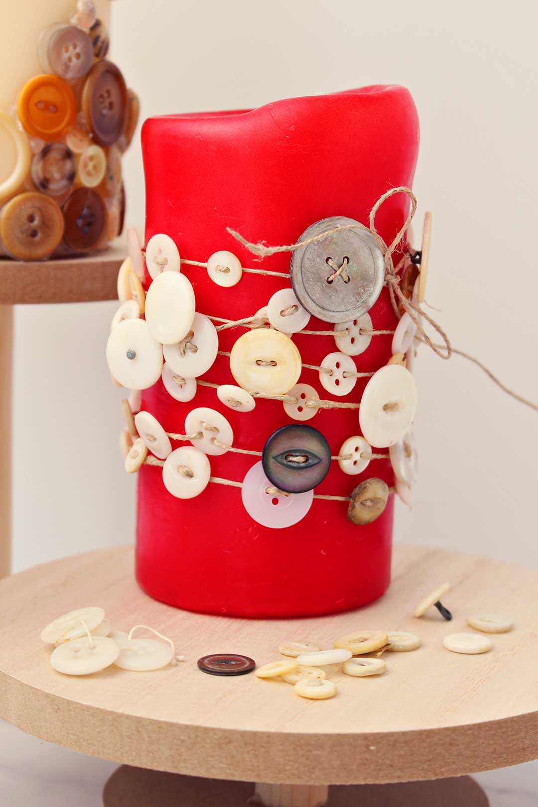 Completed red pillar candle decorated with buttons and twine resting on wooden stand.