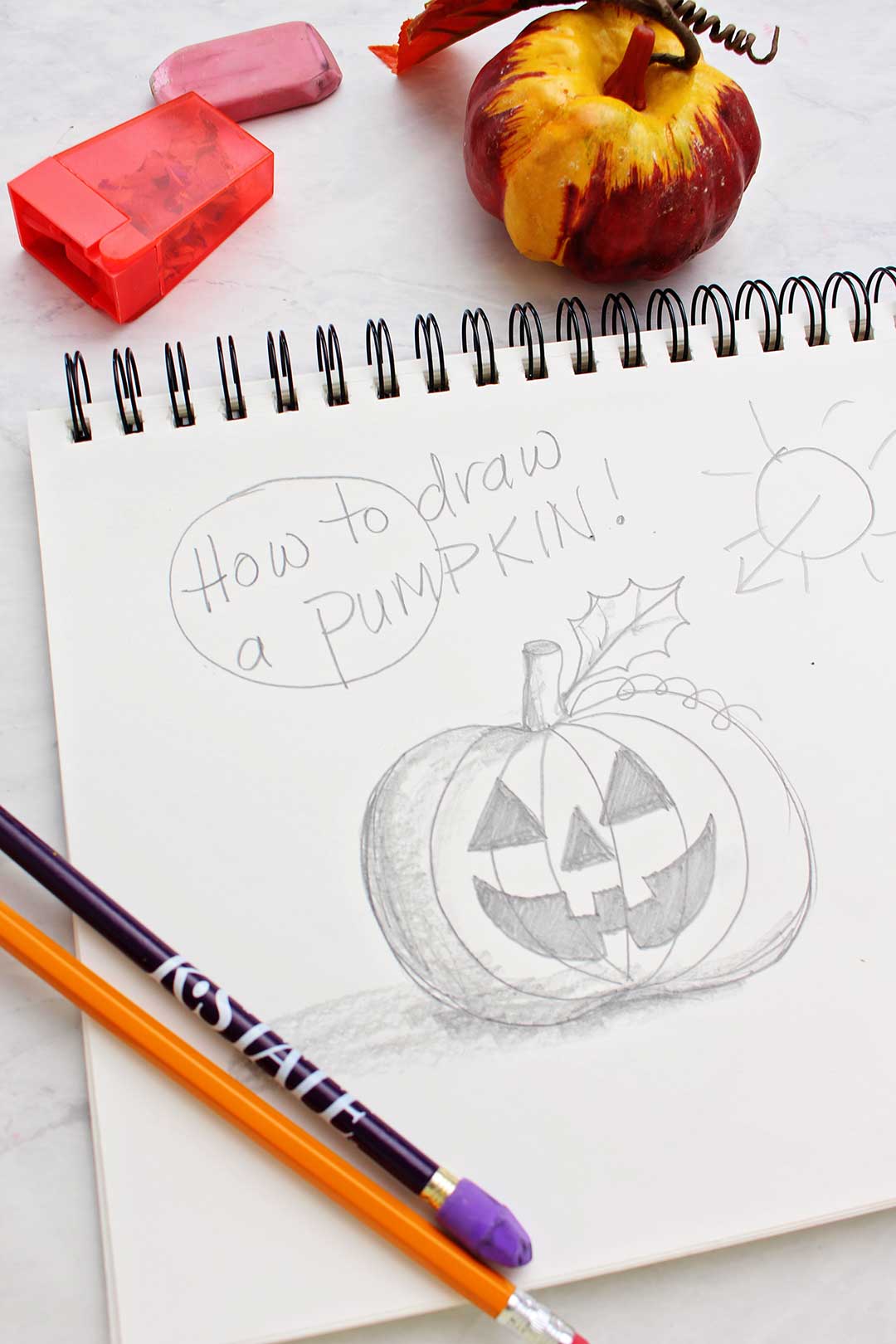 Finished pencil sketch of a pumpkin with pencils, pumpkin decoration, eraser and pencil sharpener near by.