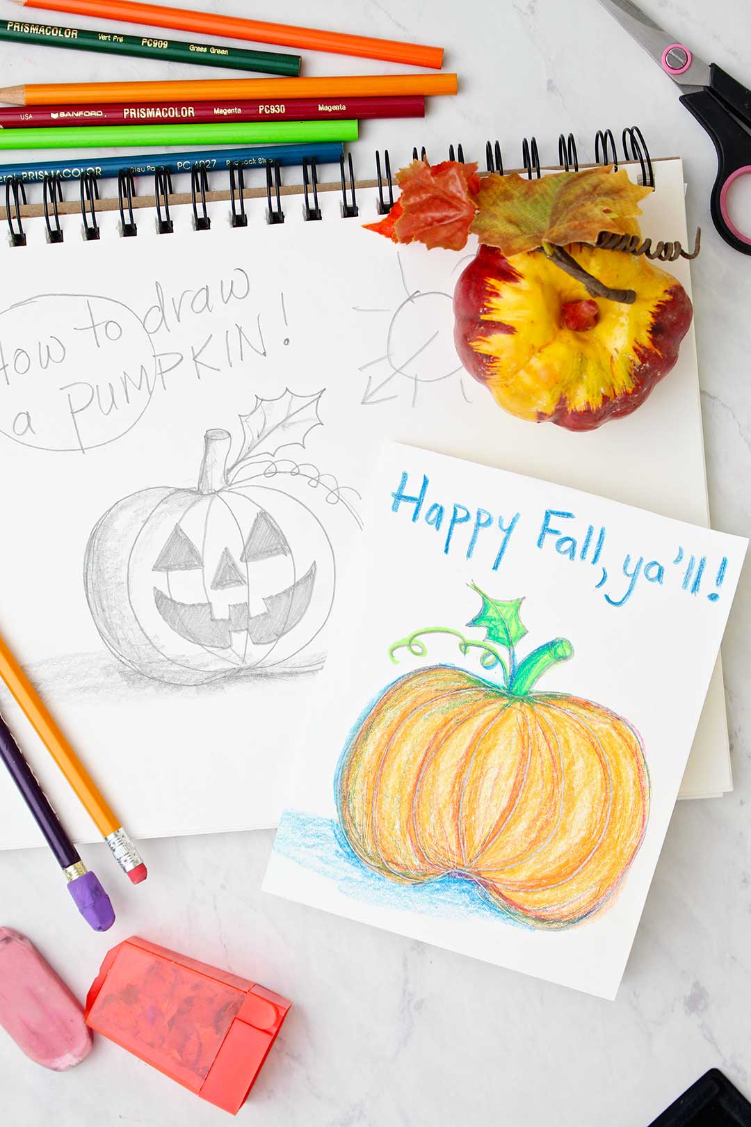 A greeting card with a colored pumpkin sketch resting on top of a jack-o-lantern pencil sketch. Drawing and coloring supplies near by.