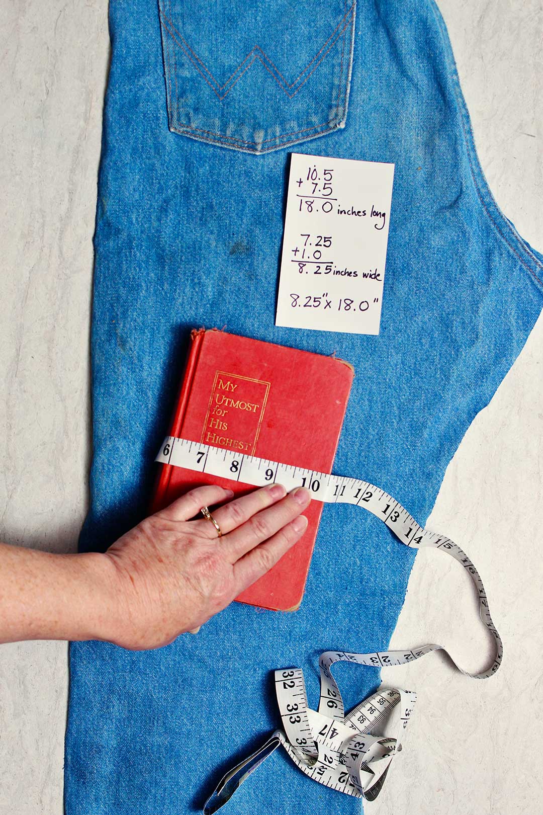 A hand measuring a book to see how much fabric will be needed. An index card showing measurements lays above it on a pair of old jeans.