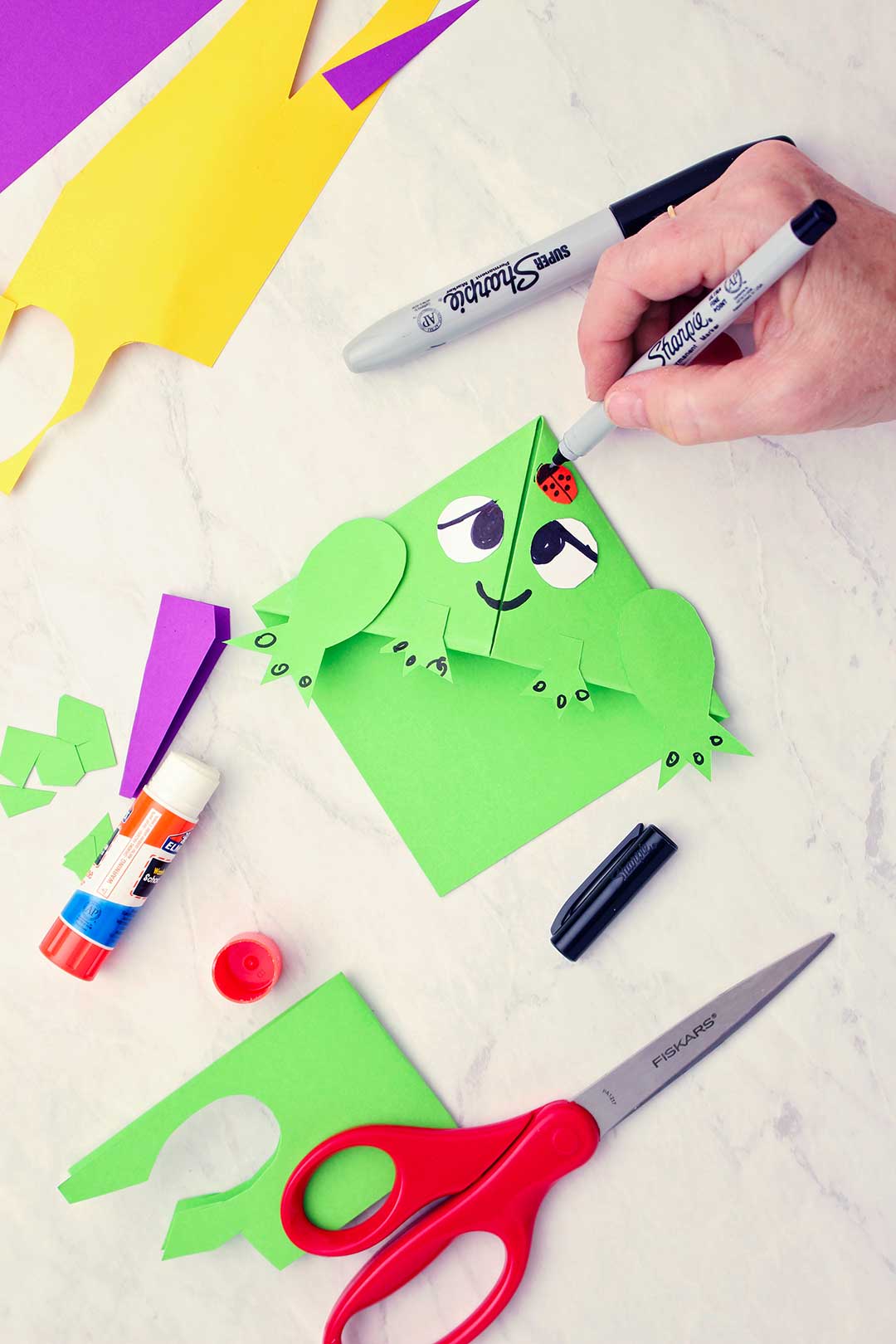 Person drawing lady bug detail on green frog origami bookmark with various supplies near by.