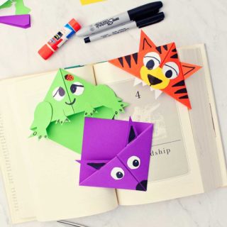 Three completed animal origami bookmarks in opened book with scraps of paper, sharpie markers and other supplies near by.