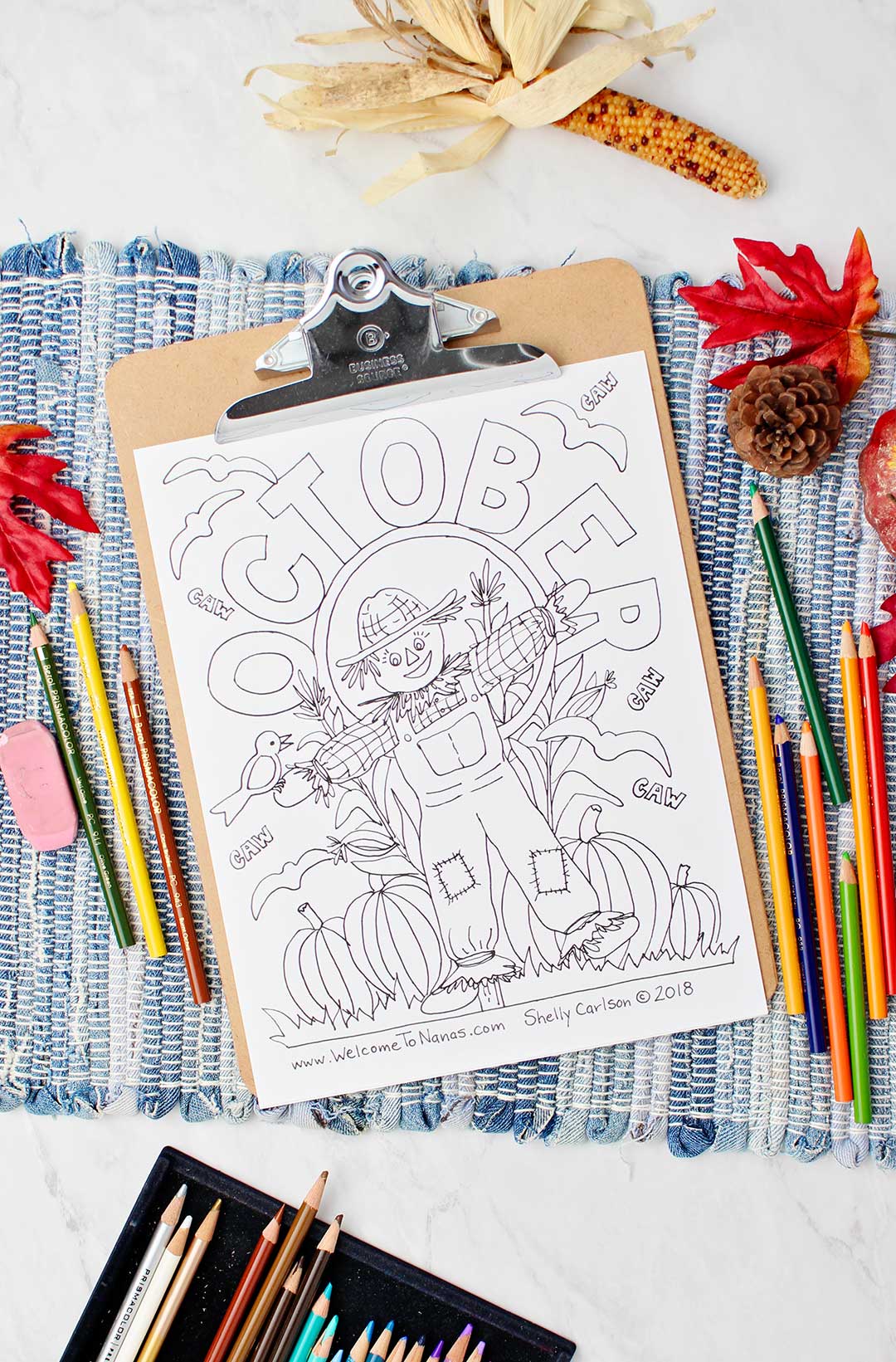 Uncolored October coloring page on blue woven placemat with colored pencils resting on it.
