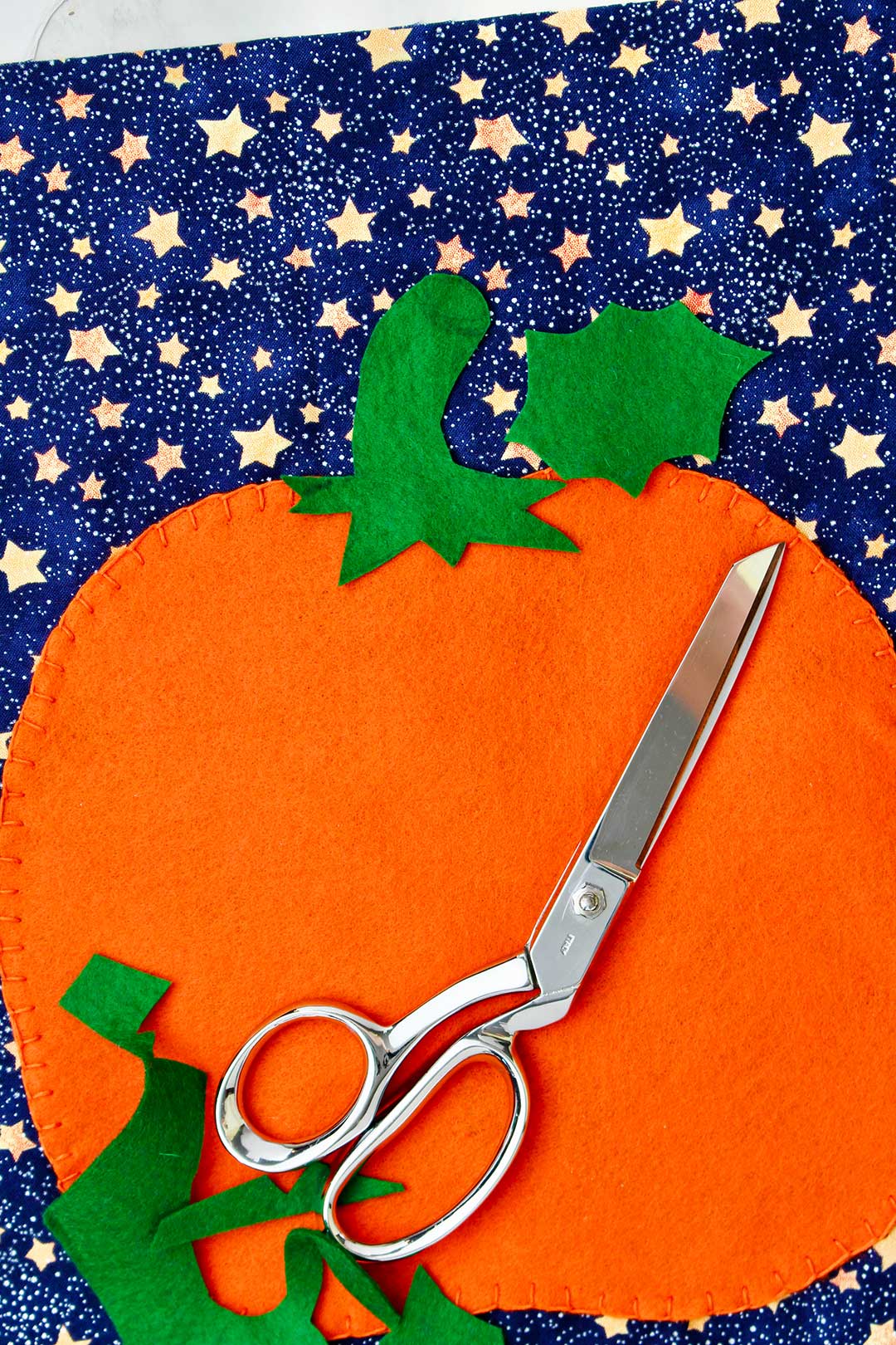Felt pumpkin cut out resting on starry blue fabric with scissors and green detail fabric scraps on top.