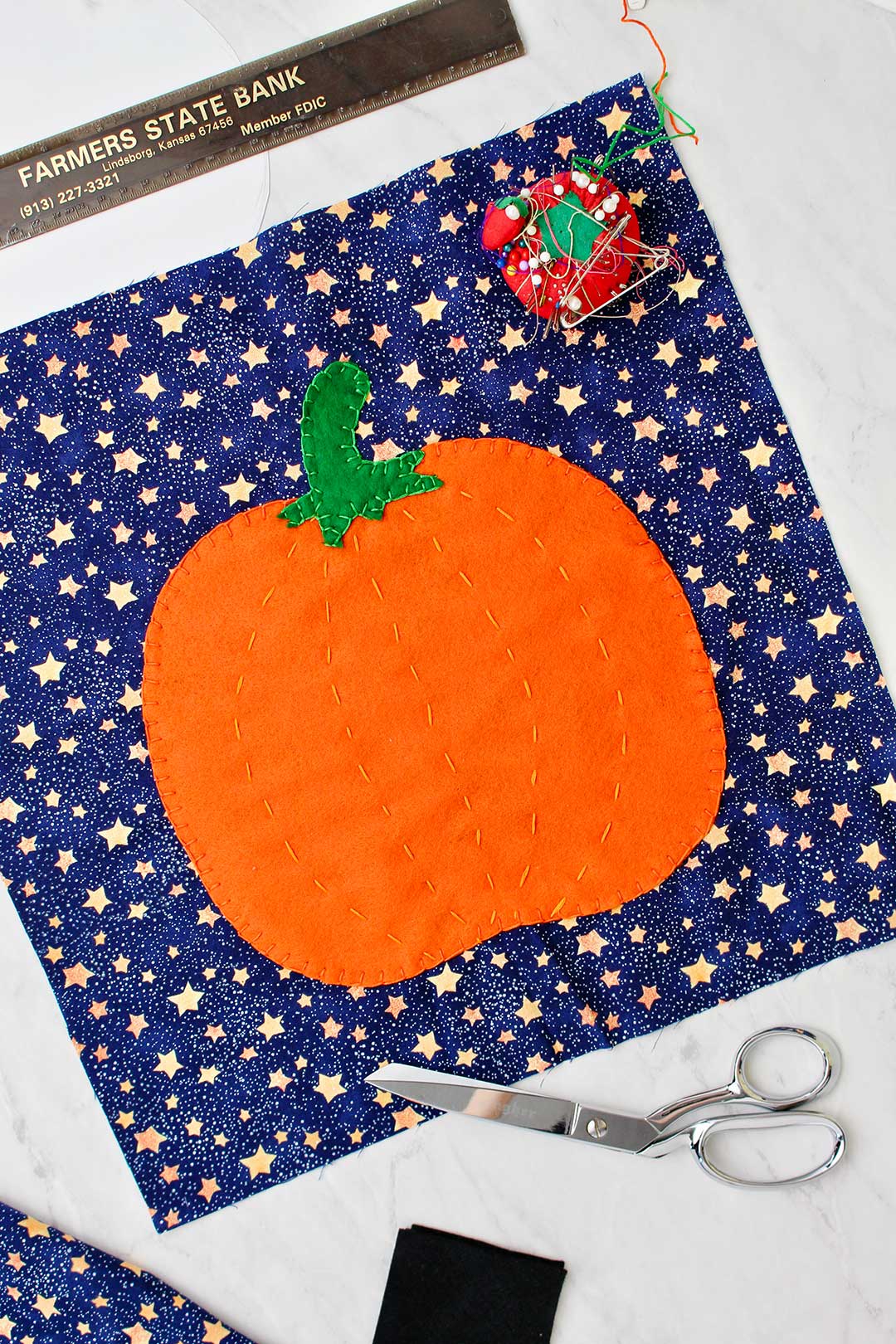 Fabric pumpkin cut out laying on a square of starry blue fabric with sewing supplies near.