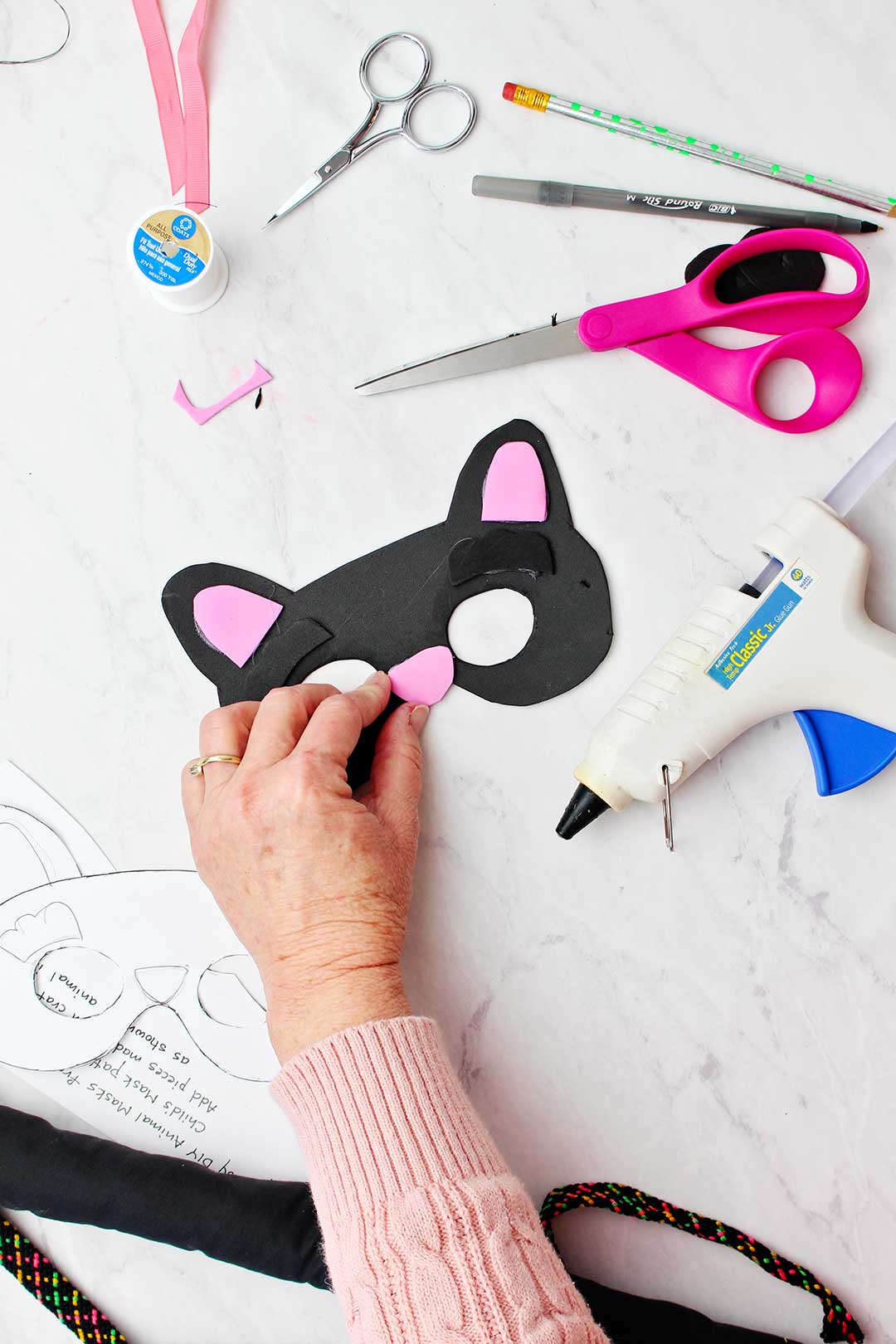 Hand hot gluing pink foam nose to black cat mask with craft supplies all around.