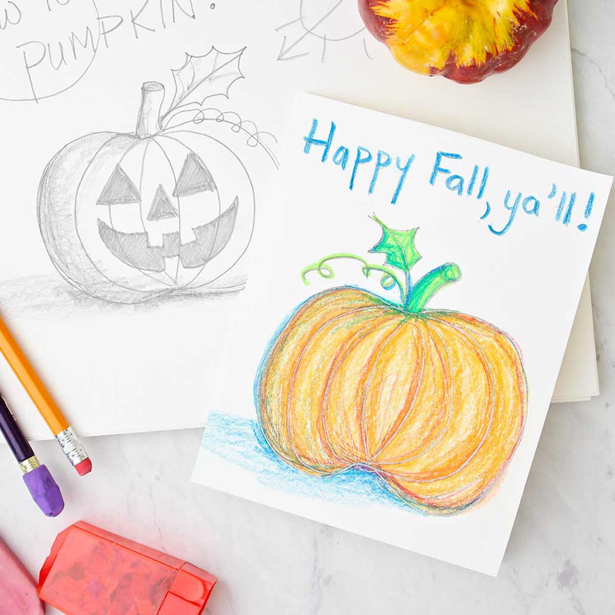 A greeting card with a colored pumpkin sketch resting on top of a jack-o-lantern pencil sketch. Drawing and coloring supplies near by.