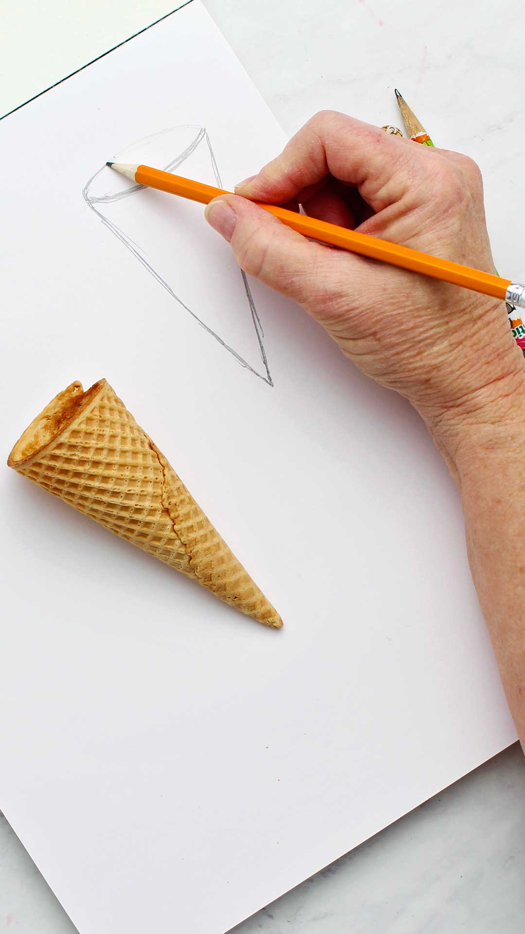 A person drawing the top oval part of the ice cream cone with an ice cream cone near by.