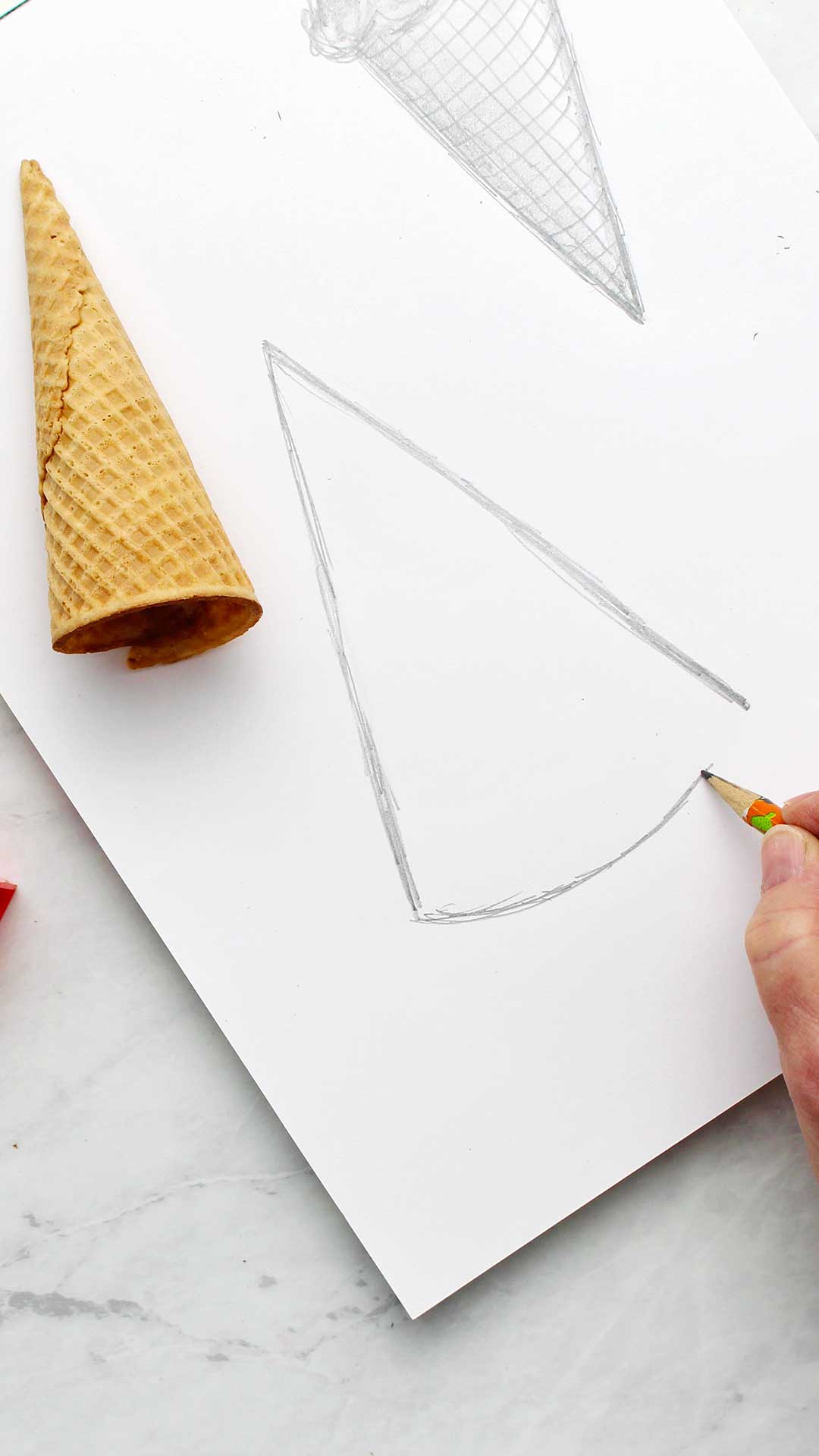 A hand sketching a cone with pencil with an ice cream cone near by as a visual aid.