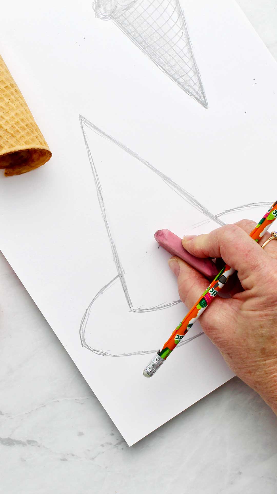 A hand erasing a line in a pencil drawing of a witches hat.