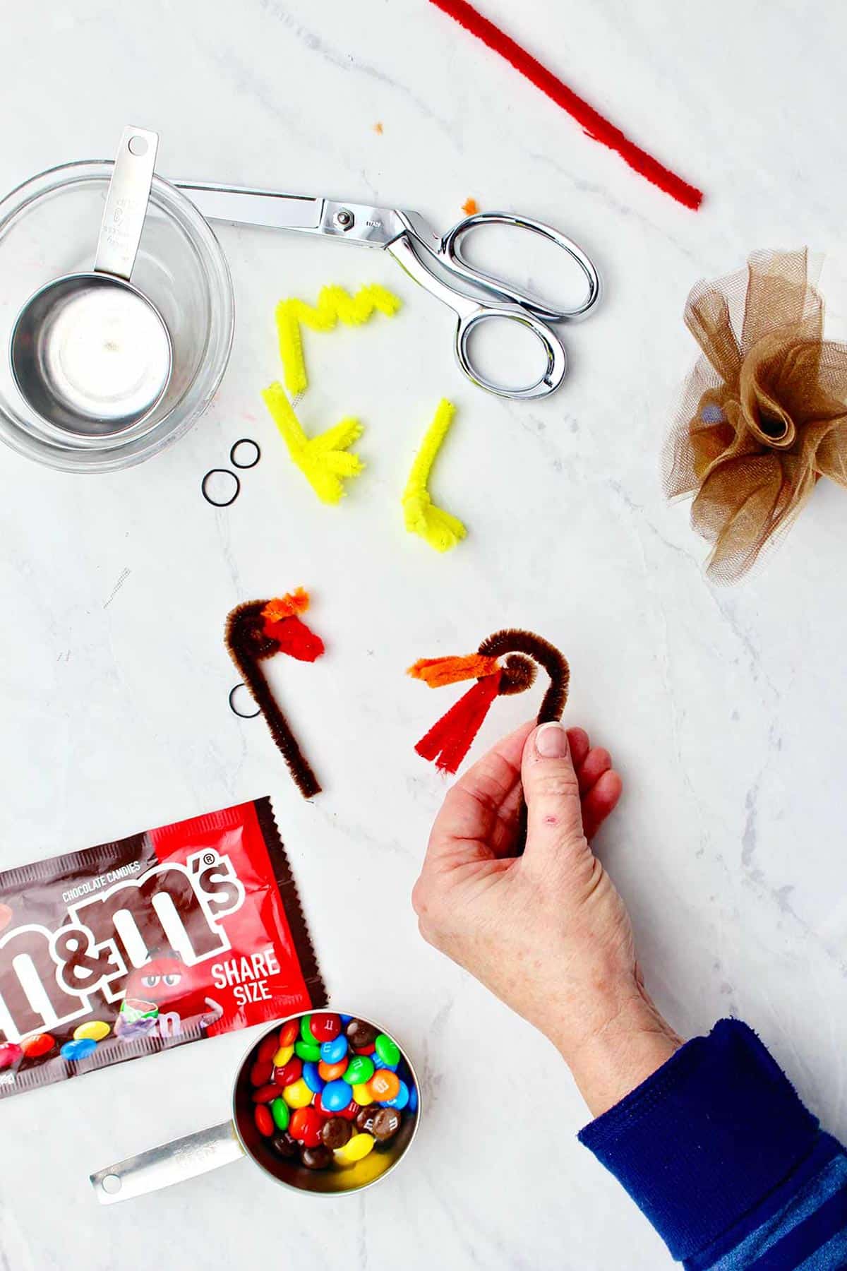 Hand holding twisted brown pipe cleaner as the start of the turkey head with various supplies near by.