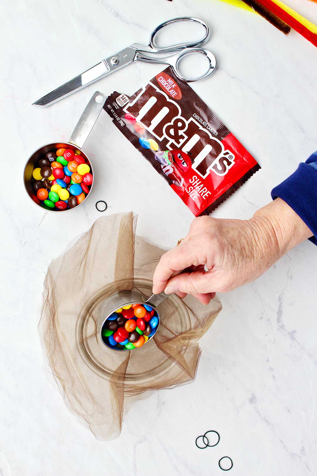 Hand pouring a measuring cup of m&ms into bowl laced with brown tulle.