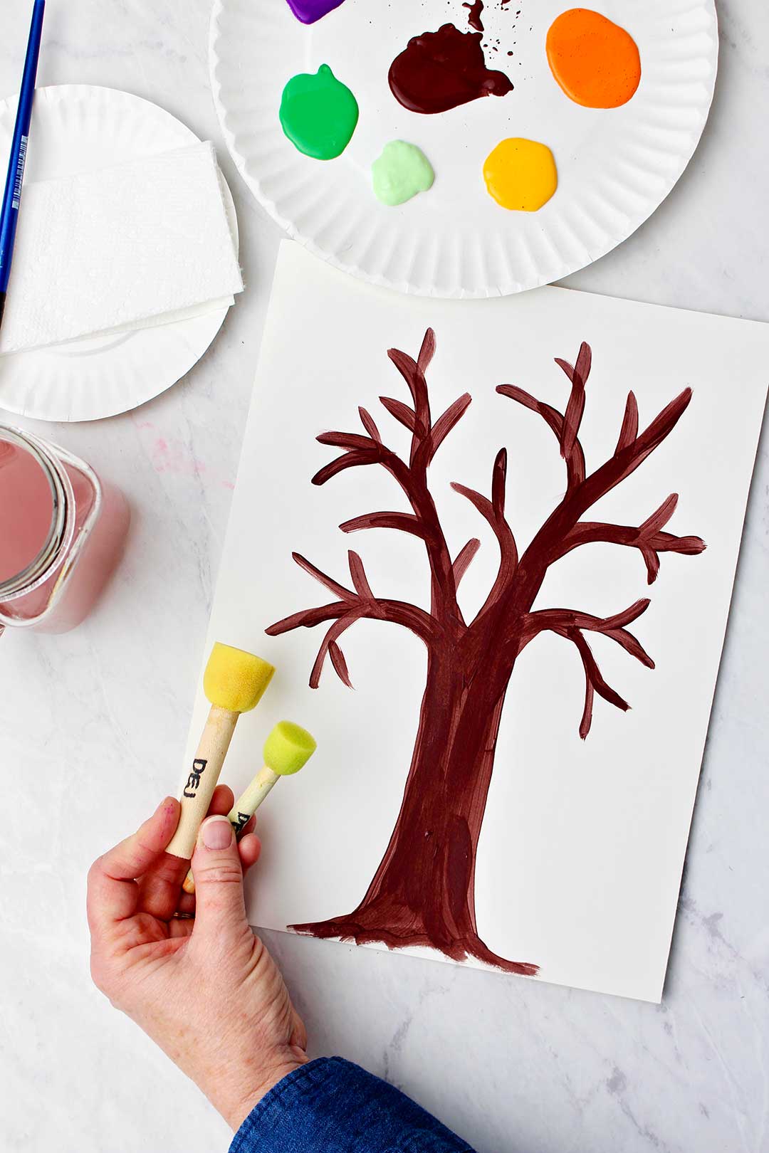 Hand showing two different circular stamping tools next to their tree trunk painting with a plate of paints and some water near by.