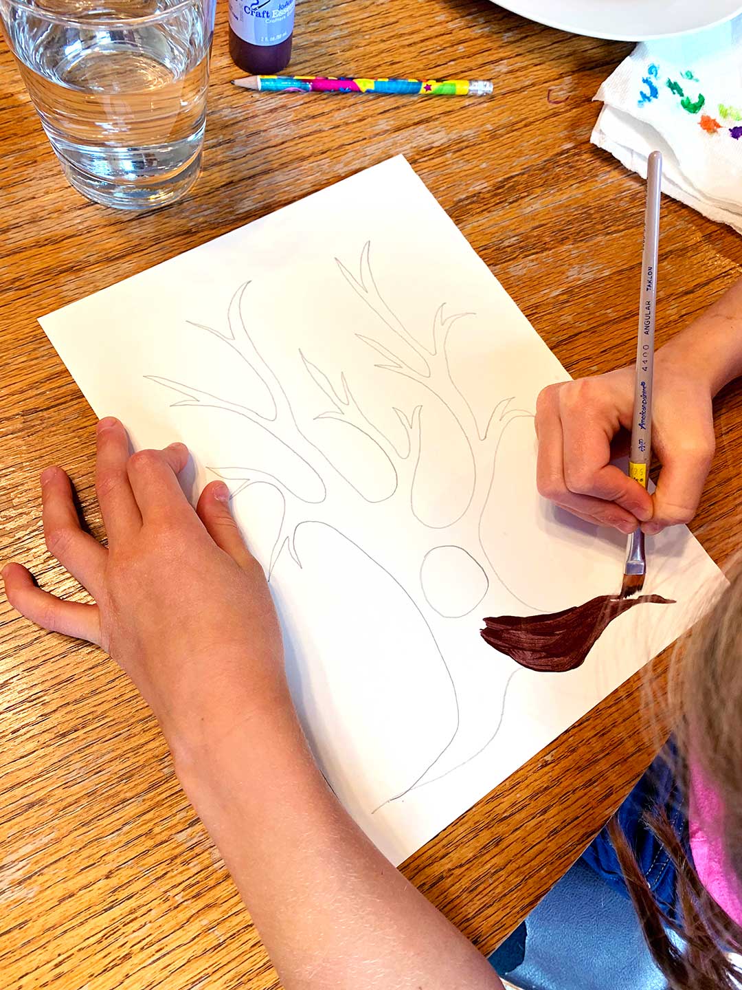 Child paints the trunk of a tree sketch at a kitchen table.
