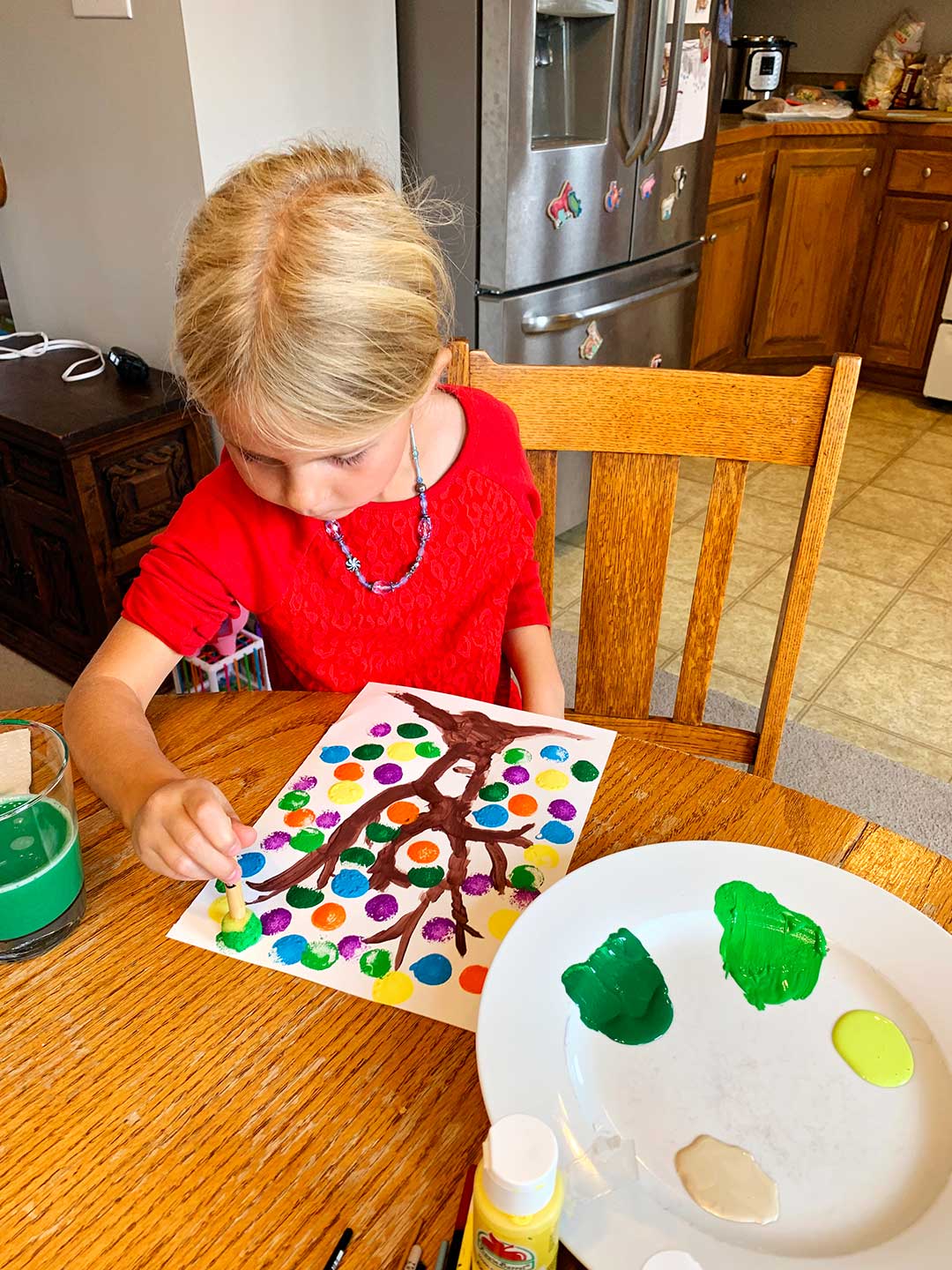 Young girl with blonde hair and red shirt stamps leaves on her tree using a circular stamp tool and green paint.