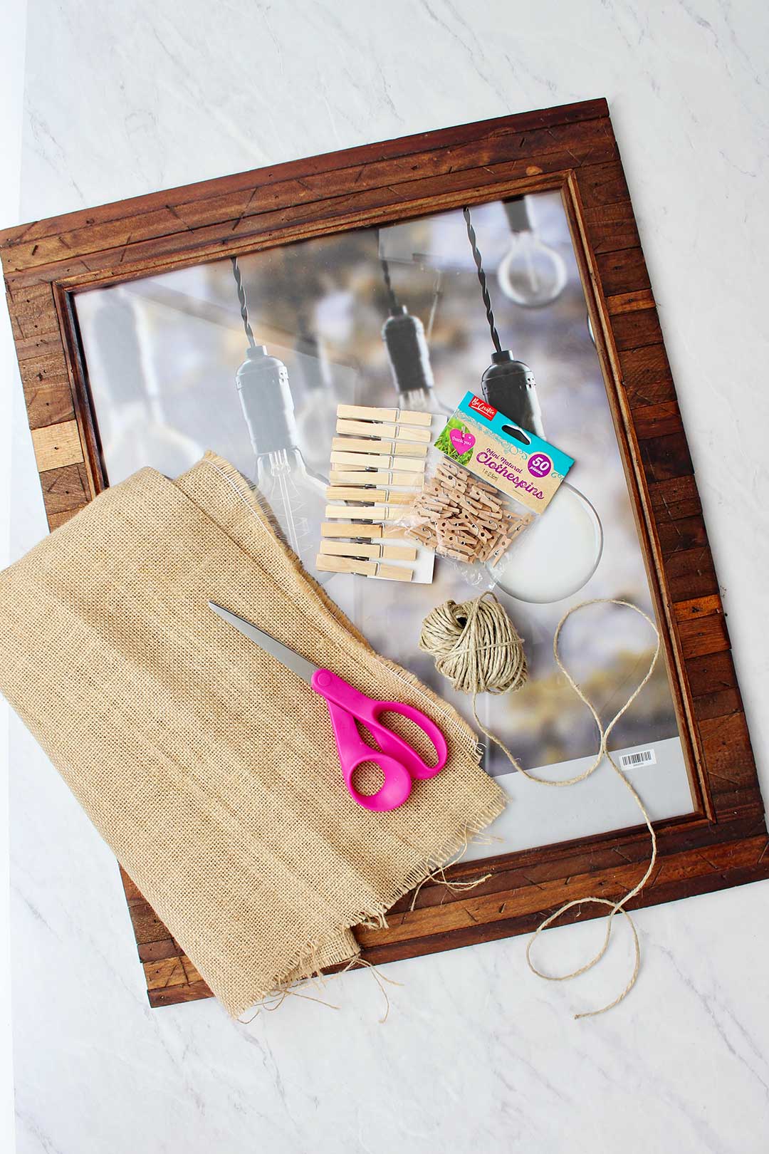 Large wooden photo frame, burlap, pink scissors, clothes pins and twine on marble counter top.