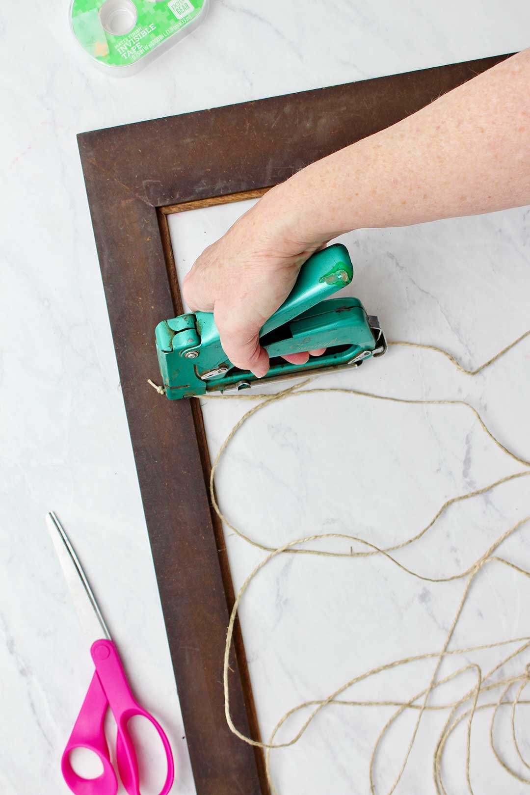 Hand attaching twine to the back of a wooden photo frame with a green staple gun with pink scissors and tape near by.