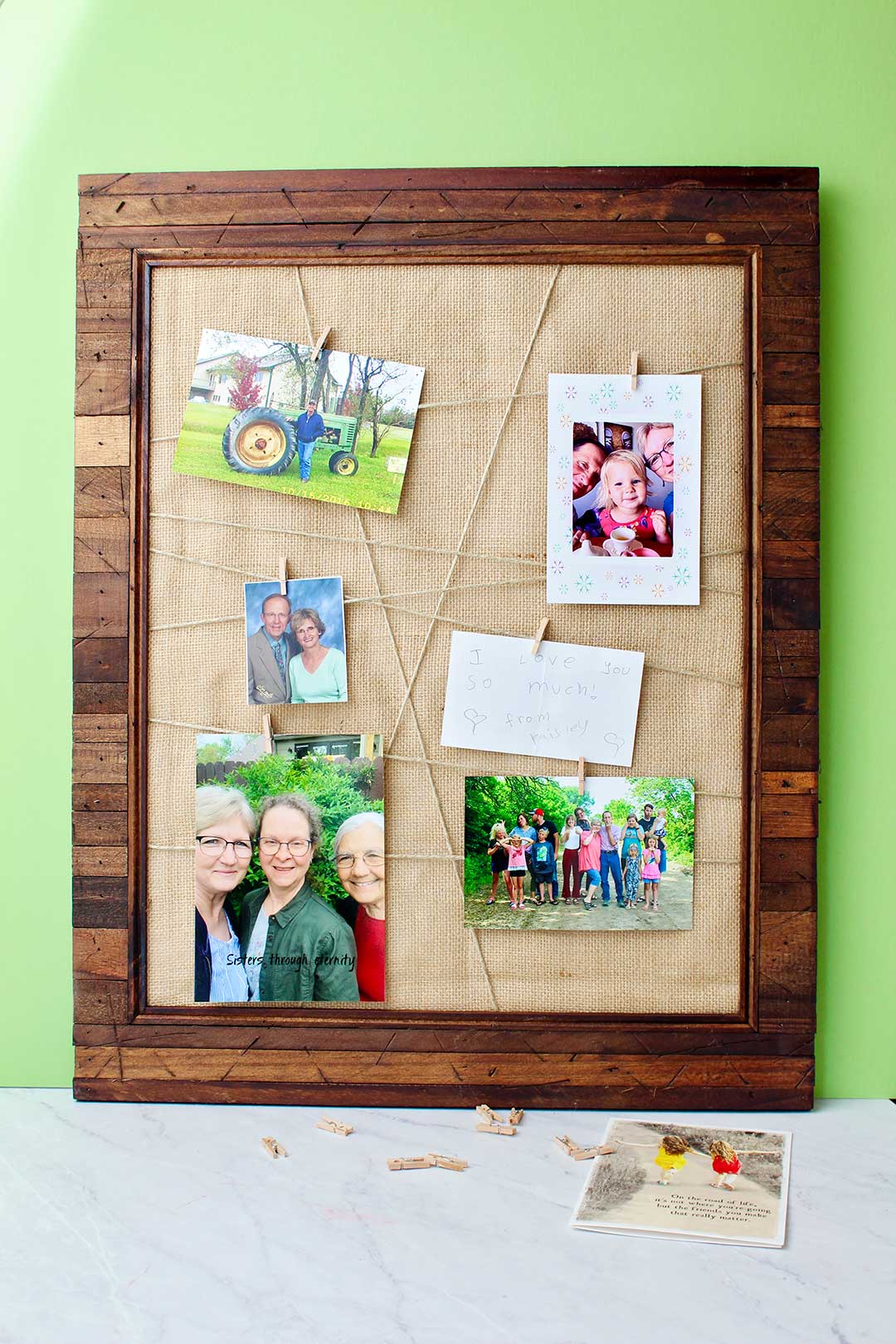 Full view of completed Upcycled Picture Frame with photos attached to criss crossed twine with clothes pins on frame with burlap backing leaning against a lime green background.