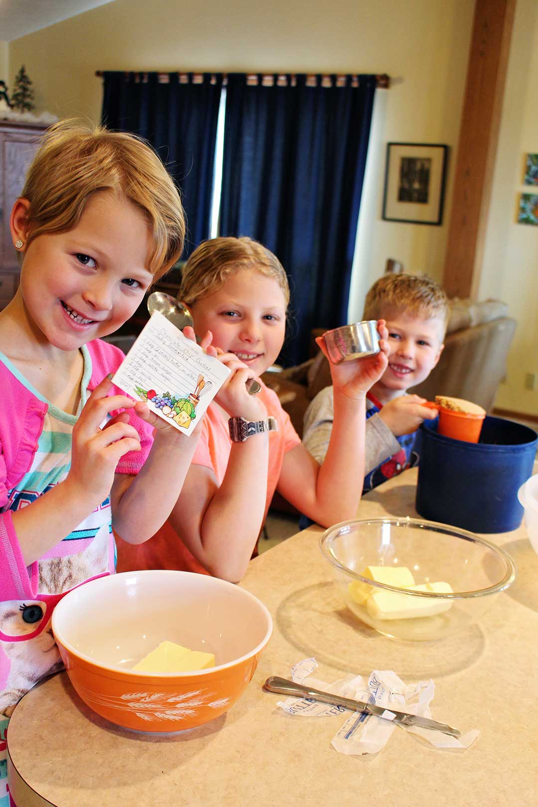 Three kids smile at the camera all holding different parts of a cookie recipe, ready to bake!