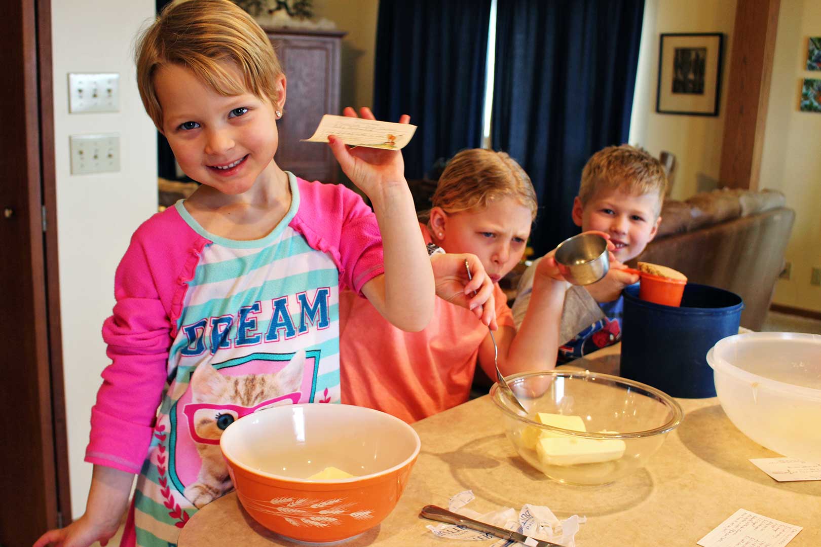 Two girls and a boy holding ingredients for a cookie recipe sitting at the kitchen counter.