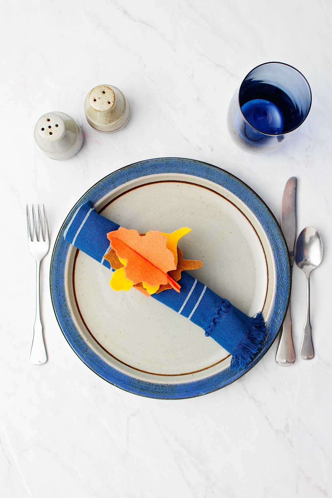 Overhead view of a table setting with plate, silverware, salt and pepper shakers, blue glass and dark blue napkin with No-Sew Felt Thanksgiving Napkin Ring holder.